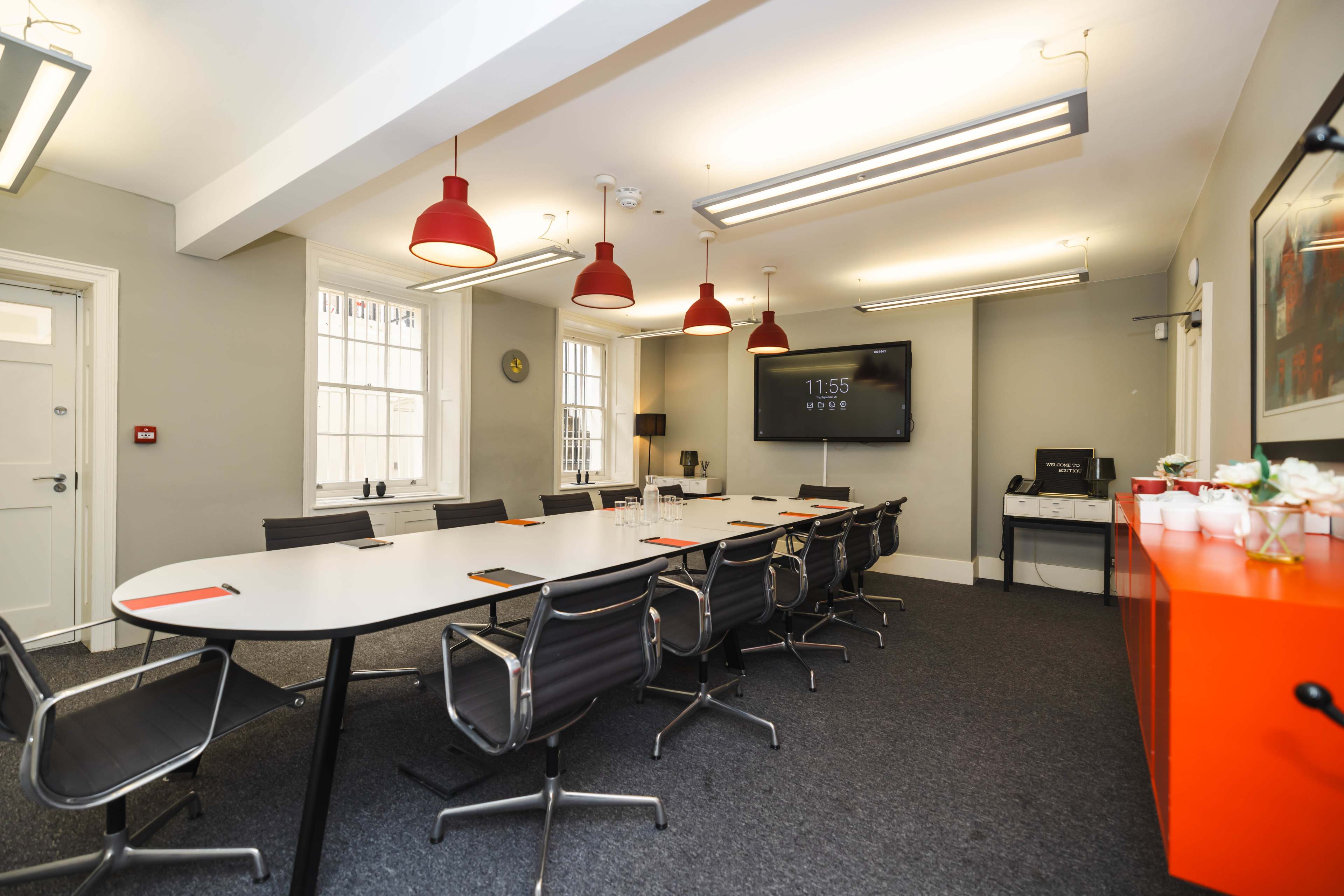 A modern conference room with a long white table, black chairs, red pendant lights, and a large screen displaying the time on one wall.