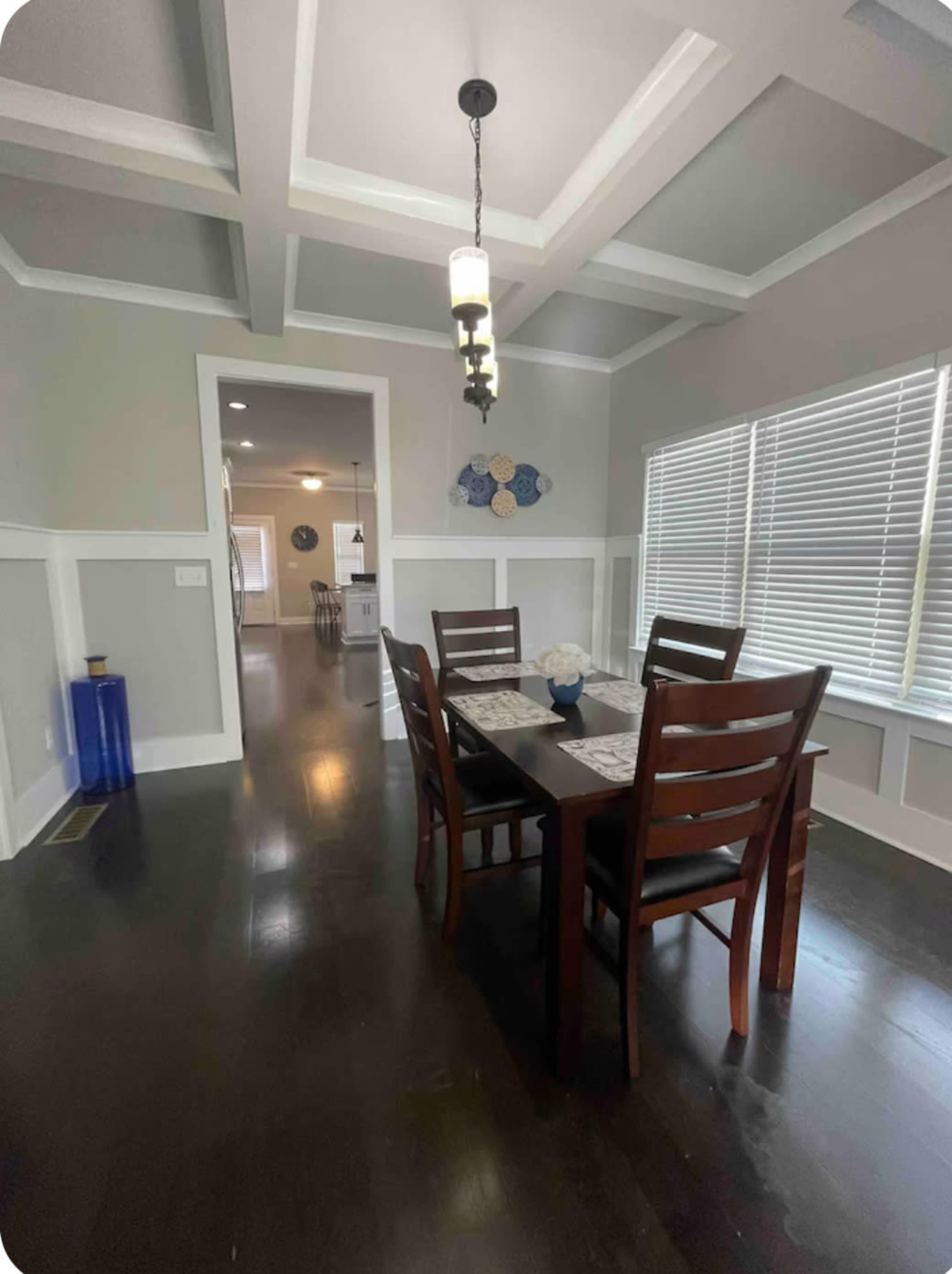 The image shows a dining area with a wooden table and chairs, a patterned centerpiece, and a window with blinds, leading to a hallway.