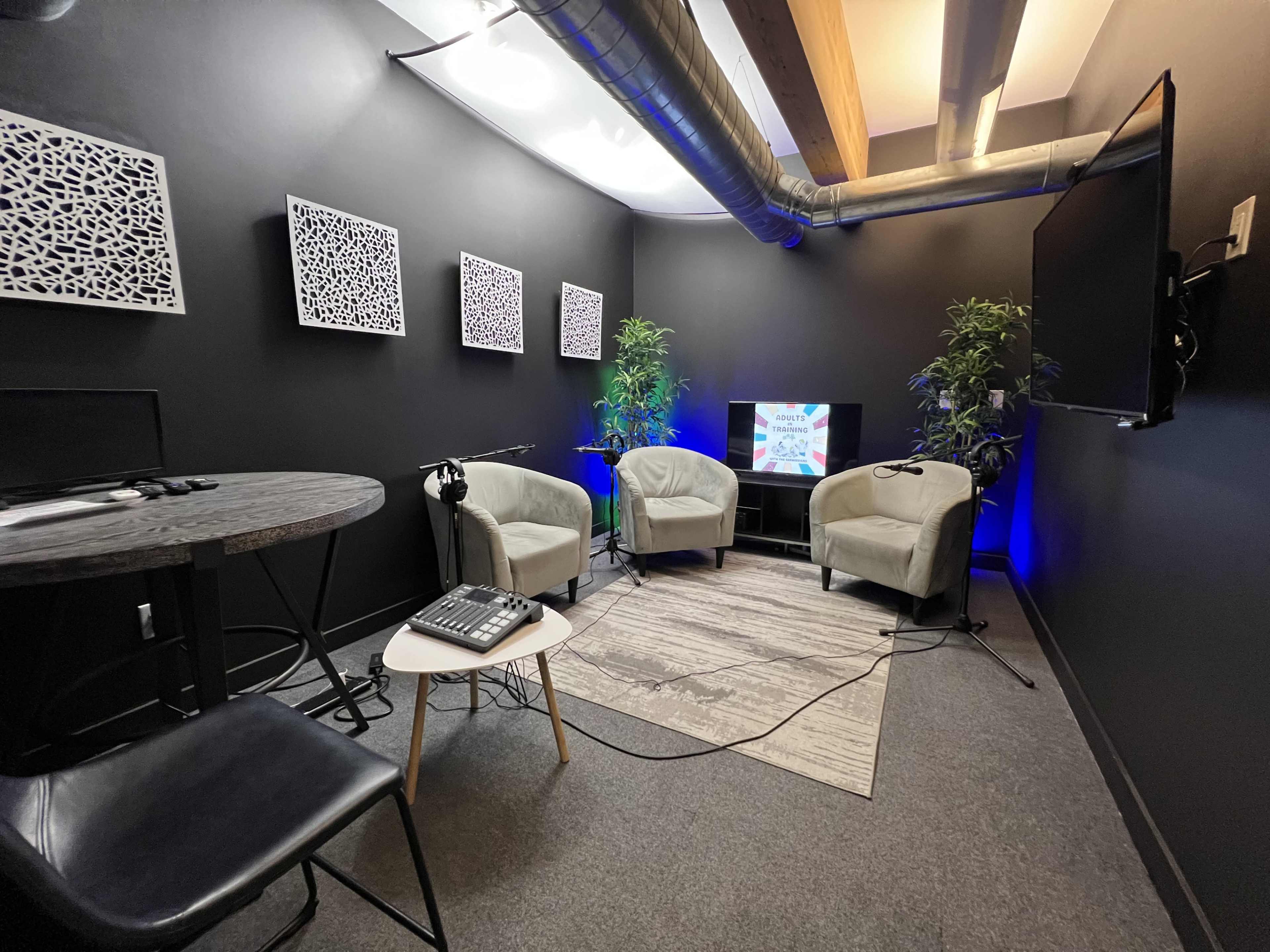 A small, modern studio space features three upholstered chairs, a table with audio equipment, and a wall-mounted TV, surrounded by dark walls and decorative wall art.