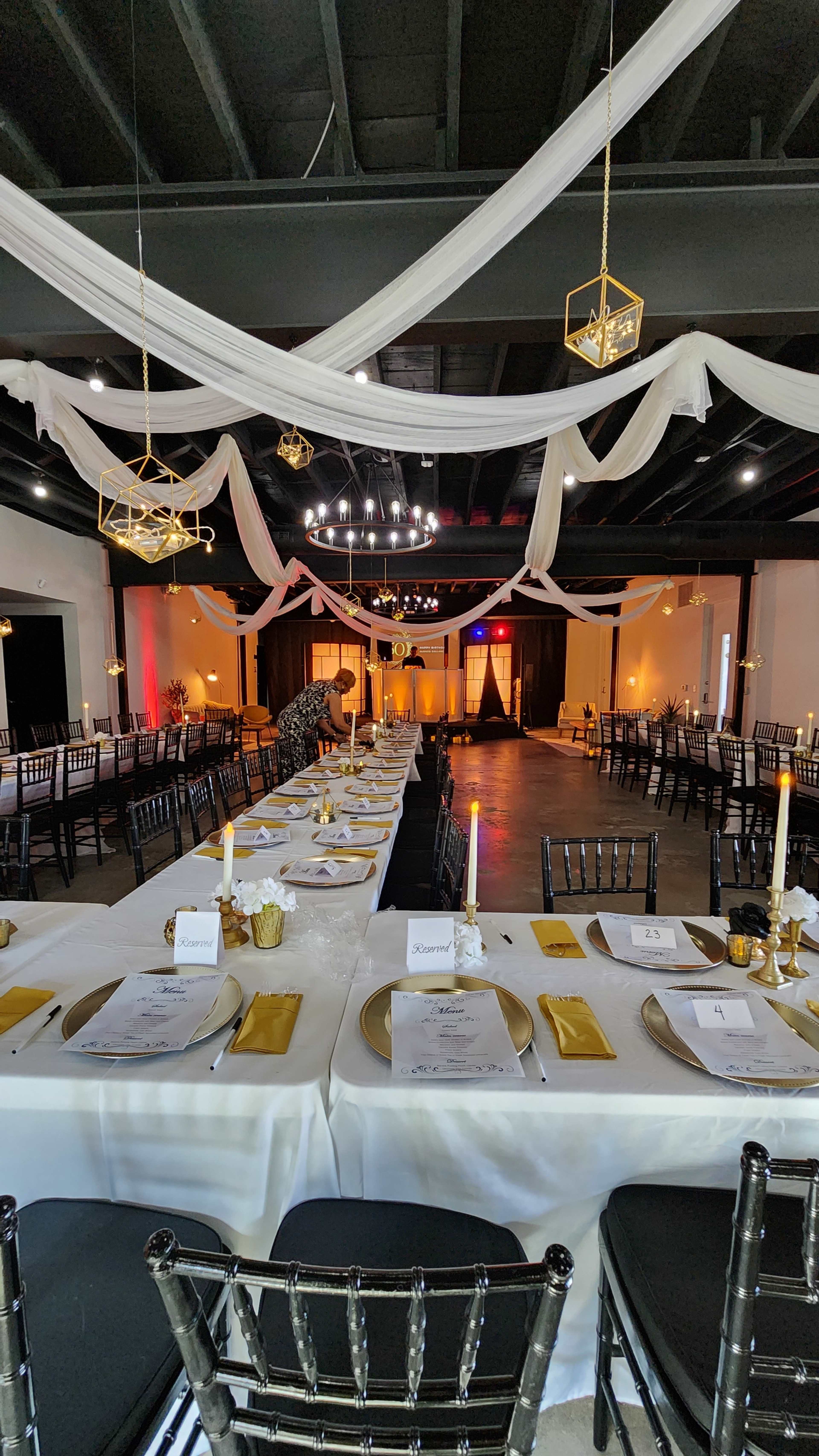 The image shows a spacious event venue set for a formal dinner, featuring long tables with white tablecloths, black chairs, and decorative elements overhead.