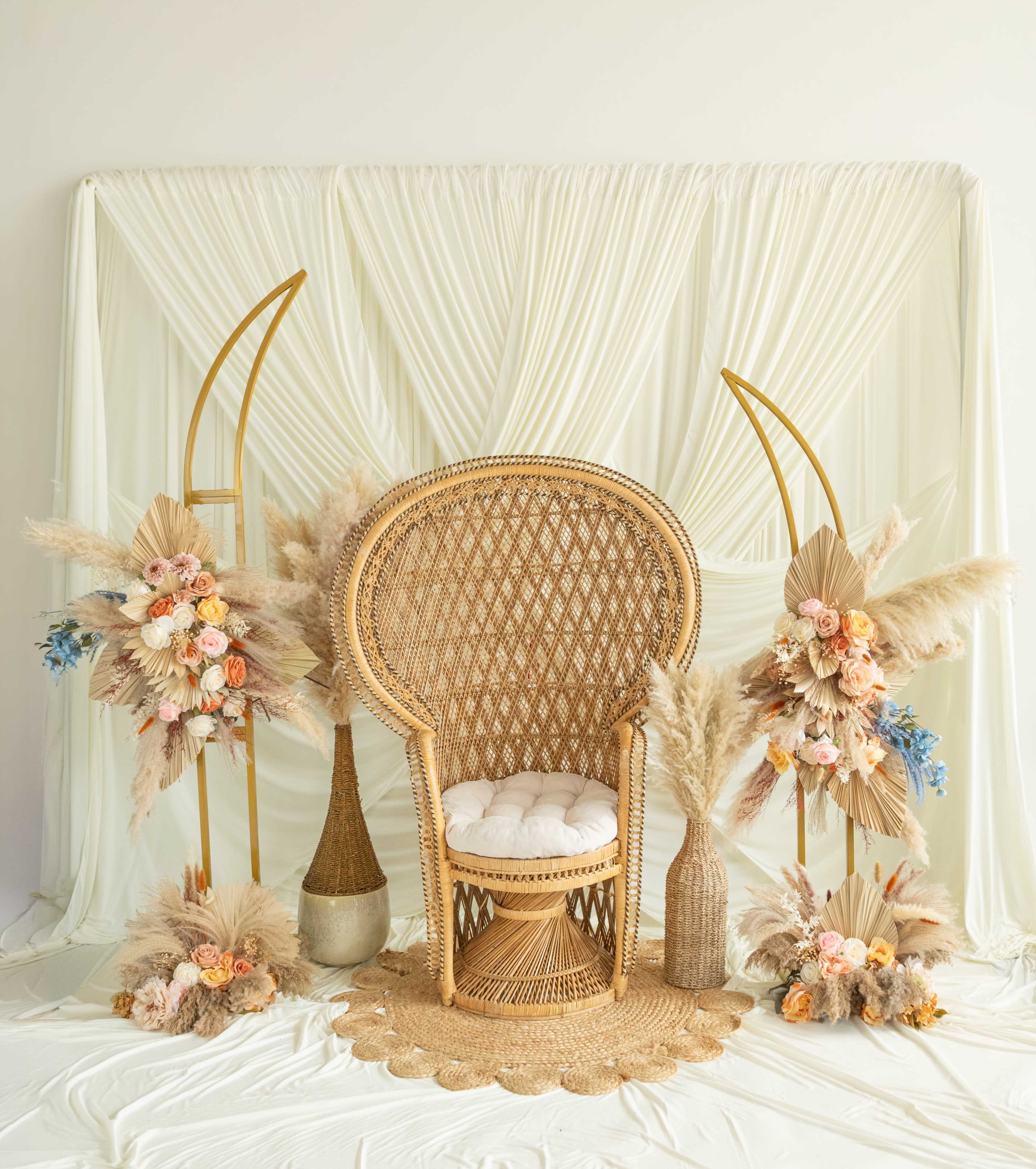 The image features a woven rattan chair surrounded by decorative floral arrangements and draped fabric in a neutral-toned setting.