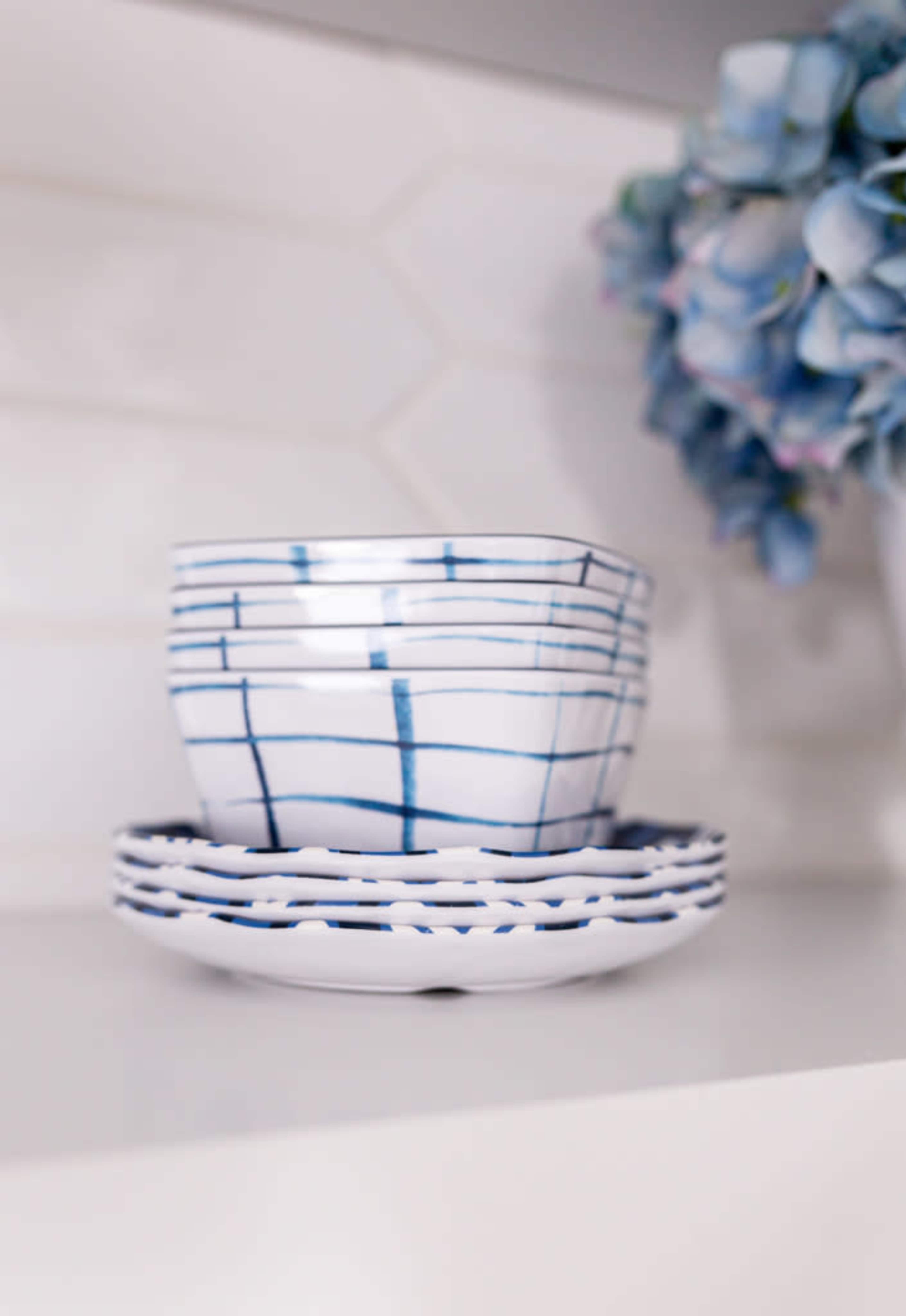 A stack of white ceramic plates and a bowl with blue geometric patterns is displayed on a shelf alongside a cluster of blue flowers.
