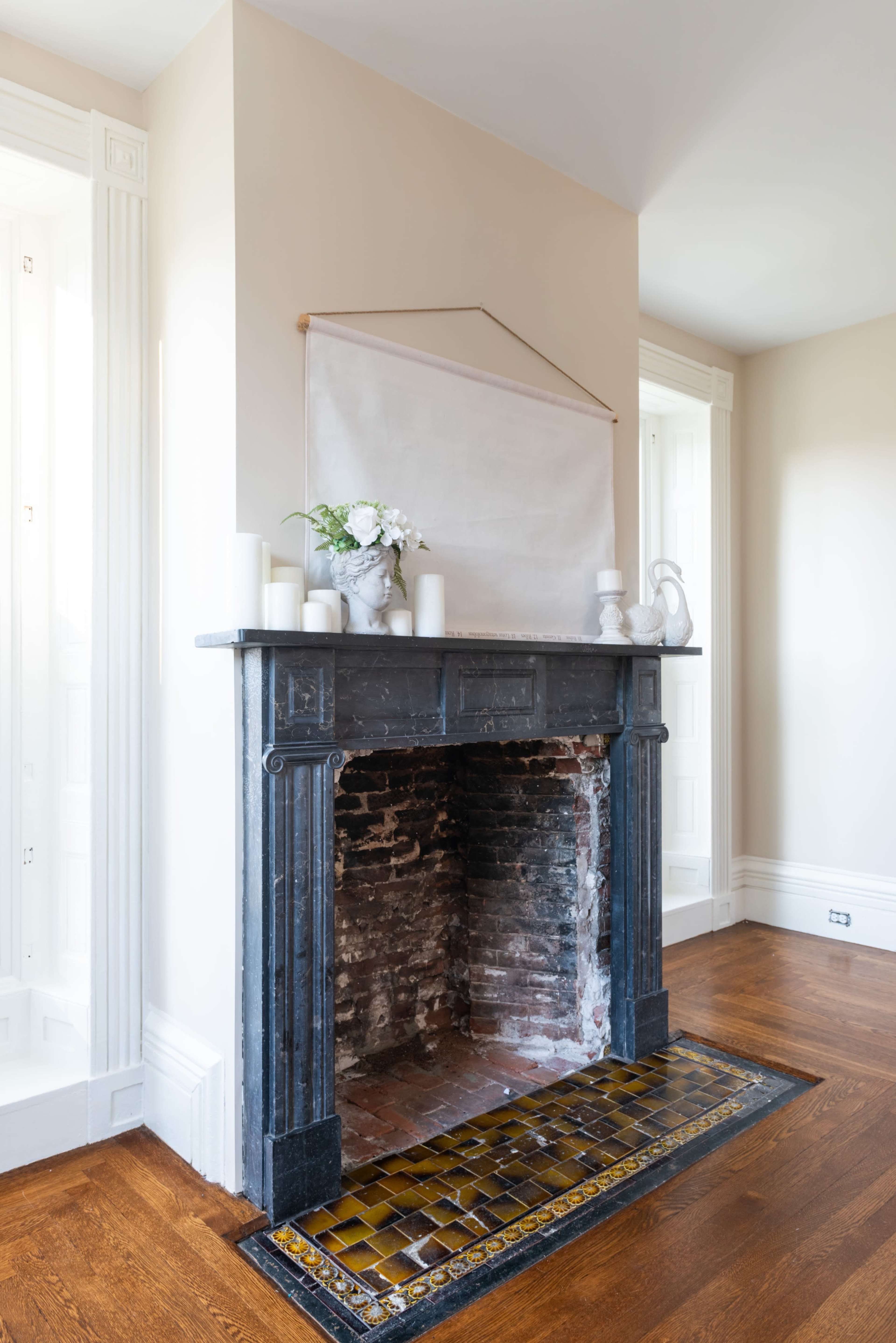 The image shows a minimalist fireplace with a black mantle and a tiled hearth, accompanied by decorative items and a blank canvas hanging above it.