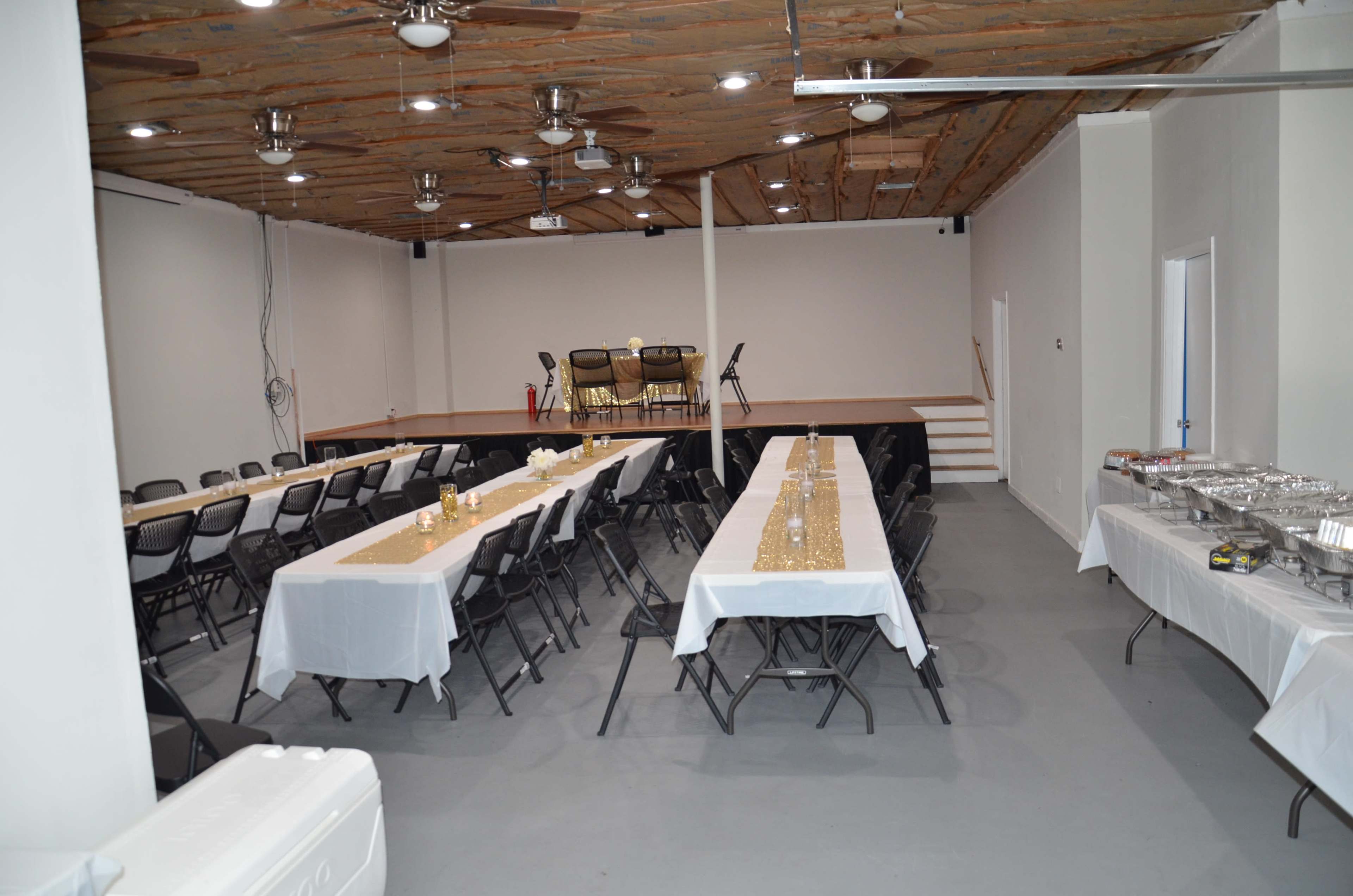 A spacious indoor event space featuring arranged tables with white tablecloths, chairs, and a small stage at the end of the room.