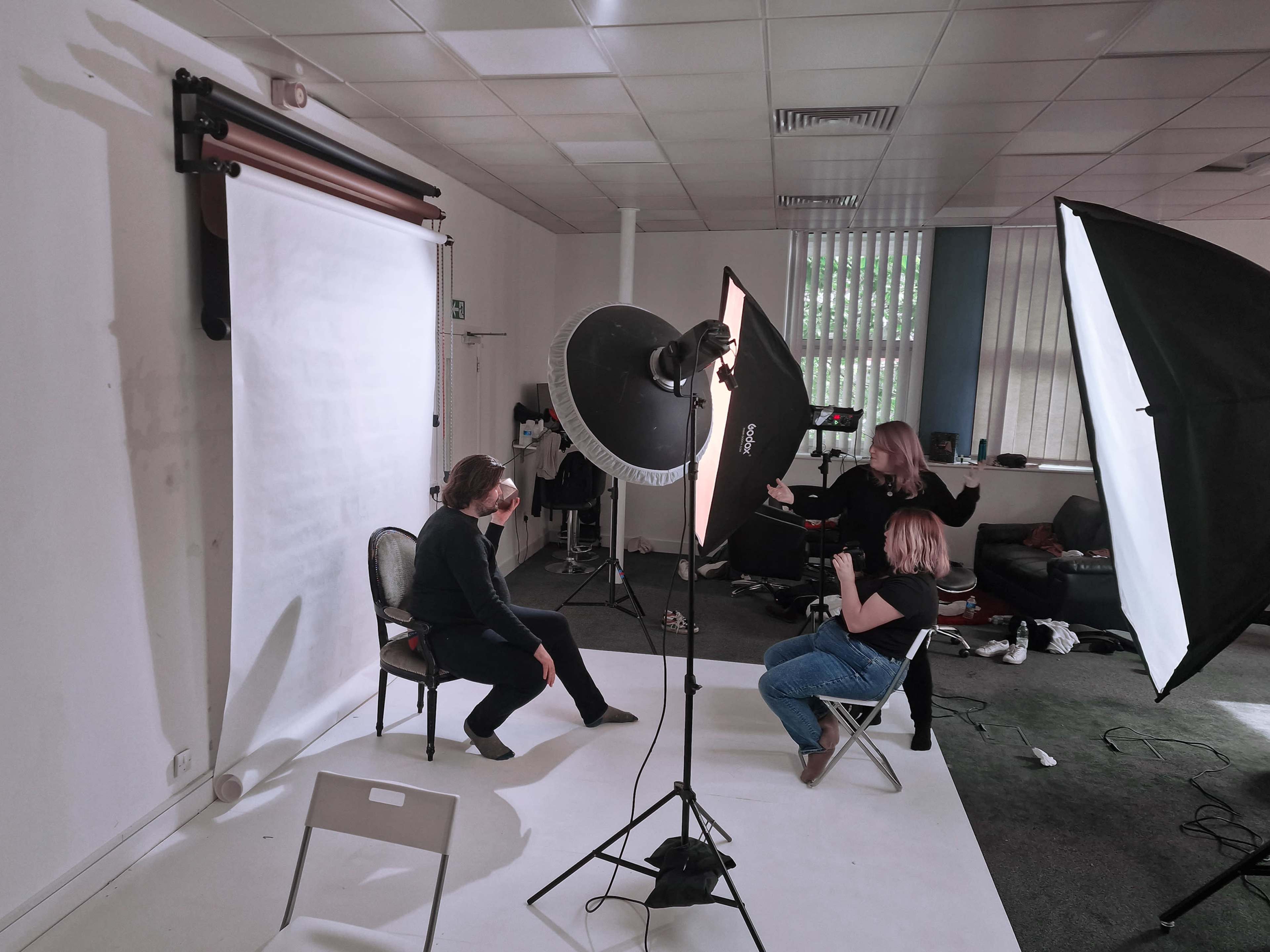 A person sits on a chair in front of a backdrop while another person photographs them, surrounded by lighting equipment in a studio.