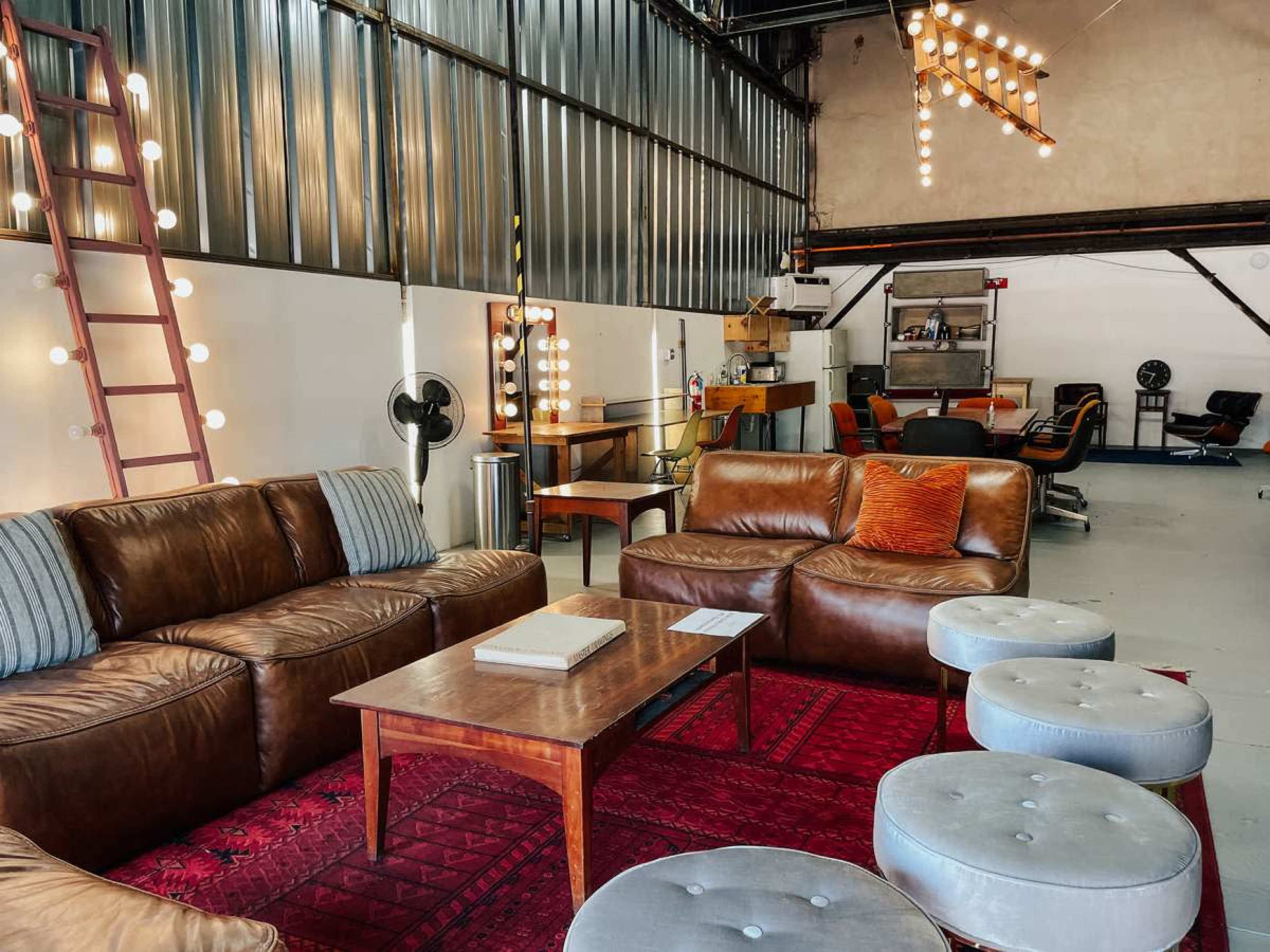 The image shows a modern industrial space with a lounge area featuring brown leather sofas, a wooden coffee table, and round upholstered stools, along with a work area in the background equipped with office chairs and desks.
