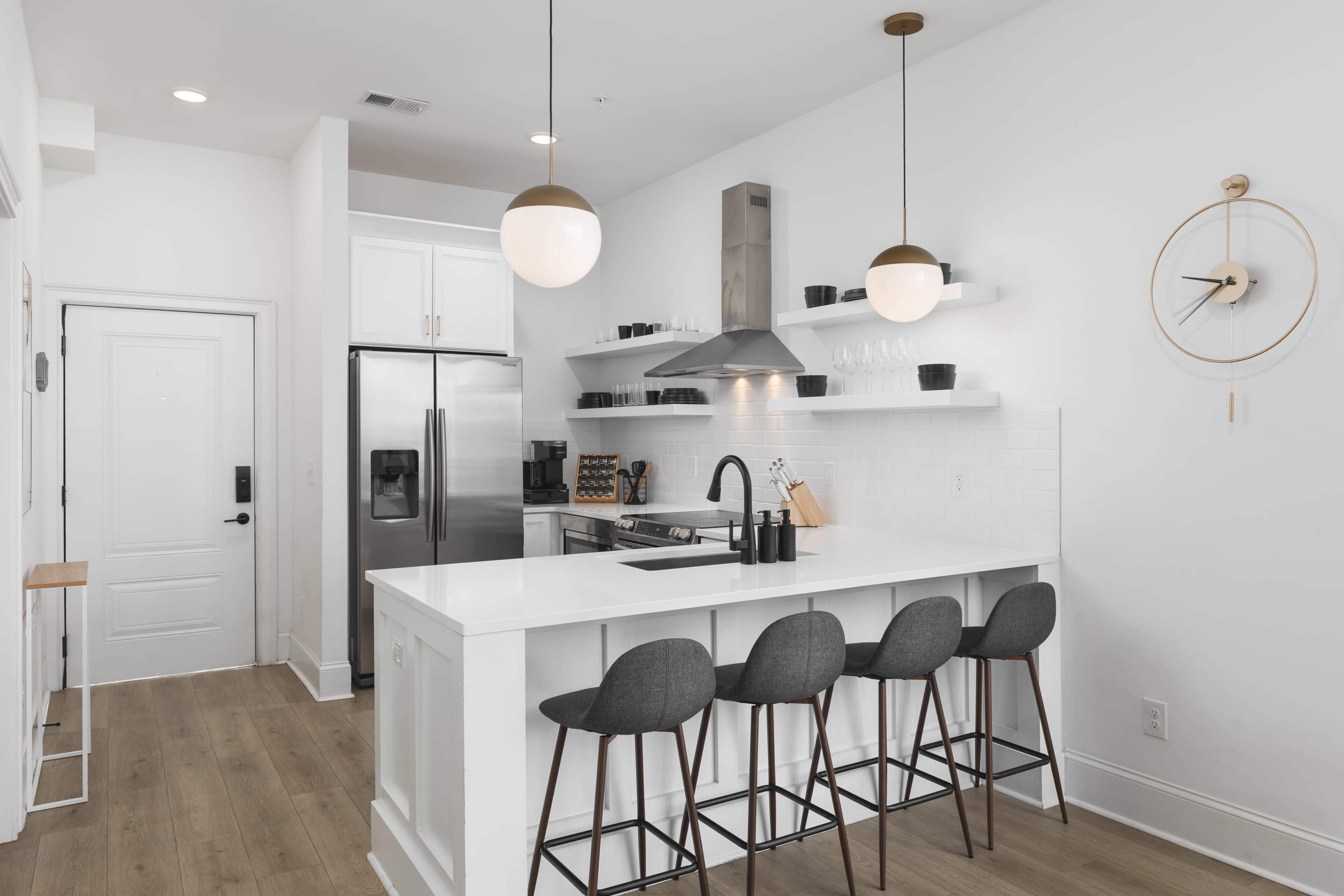 A modern kitchen features a white countertop island with four gray stools, stainless steel appliances, and open shelves displaying kitchenware against a bright wall.