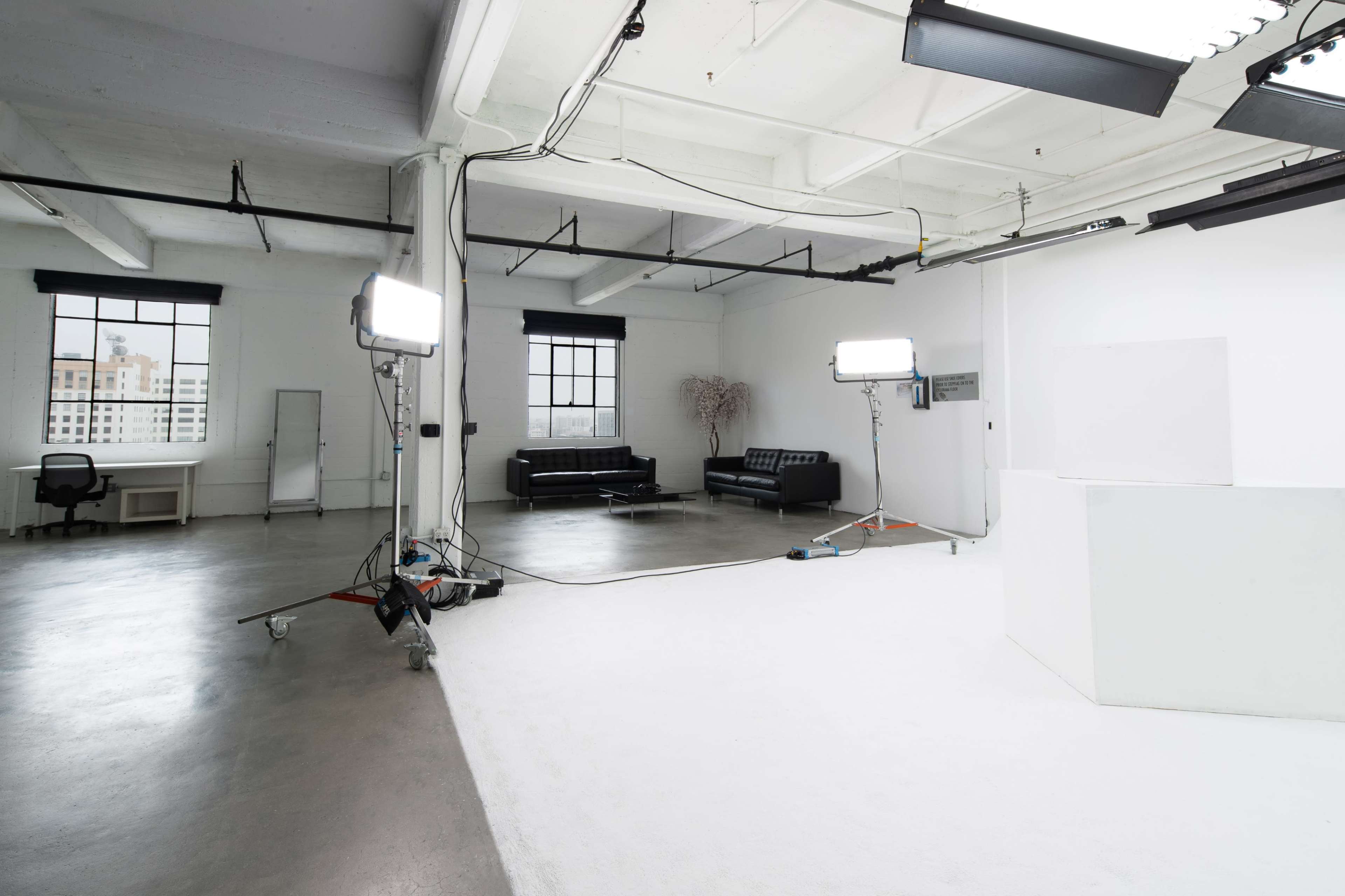The image shows a spacious, well-lit studio with white walls, a white floor, a black couch, and multiple lights set up for photography.