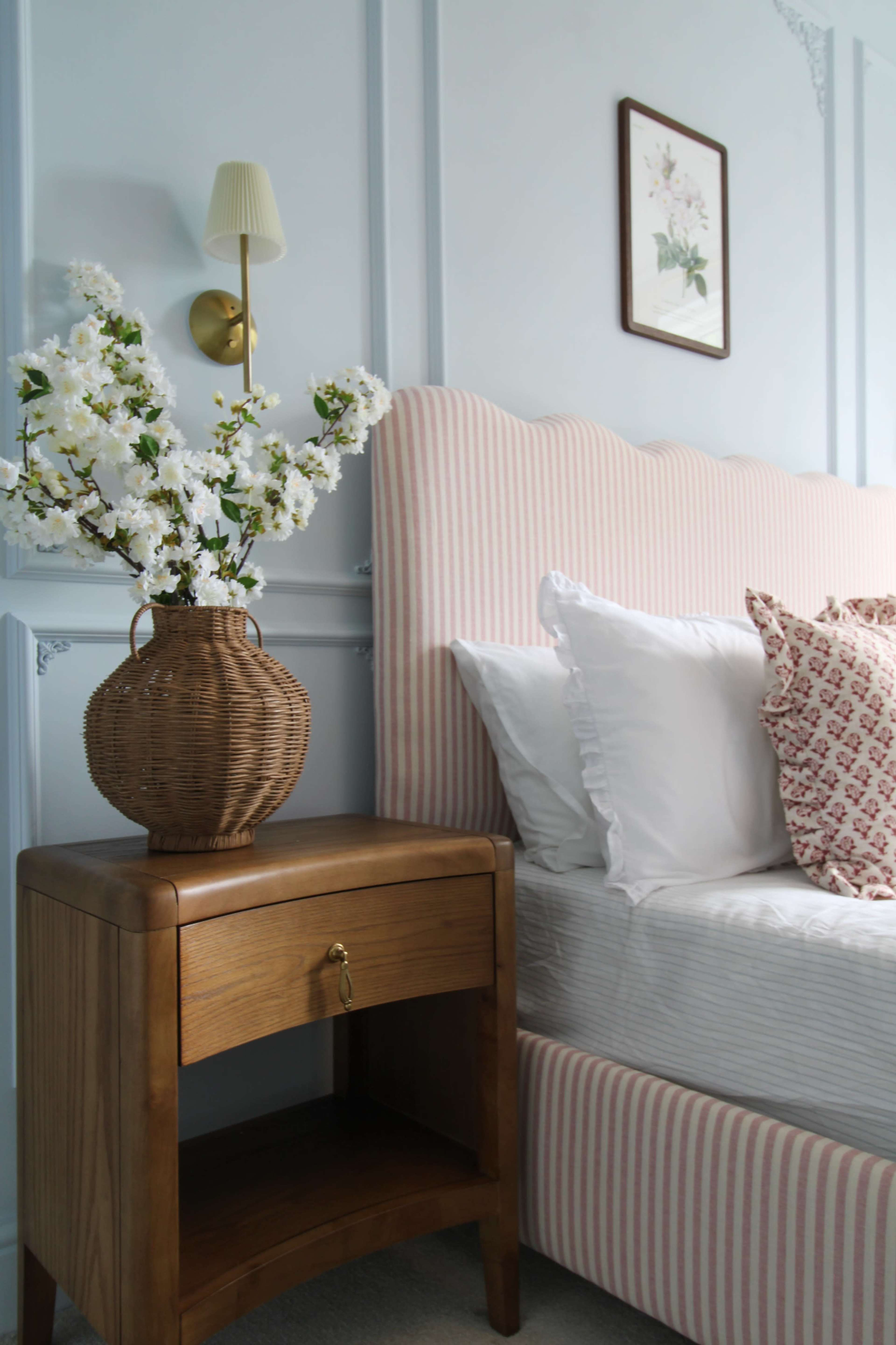 A bedside table with a wicker vase of flowers stands next to a striped upholstered headboard and neatly made bed.