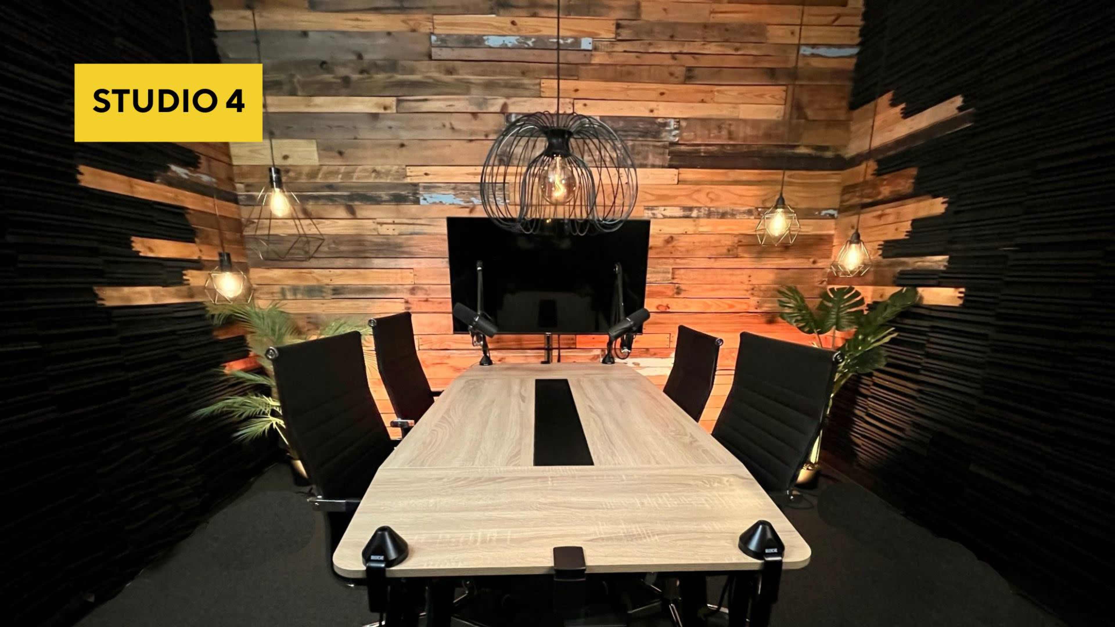 A modern conference room features a large wooden table surrounded by black chairs, with a television on the wall and pendant lights hanging from a wooden backdrop.