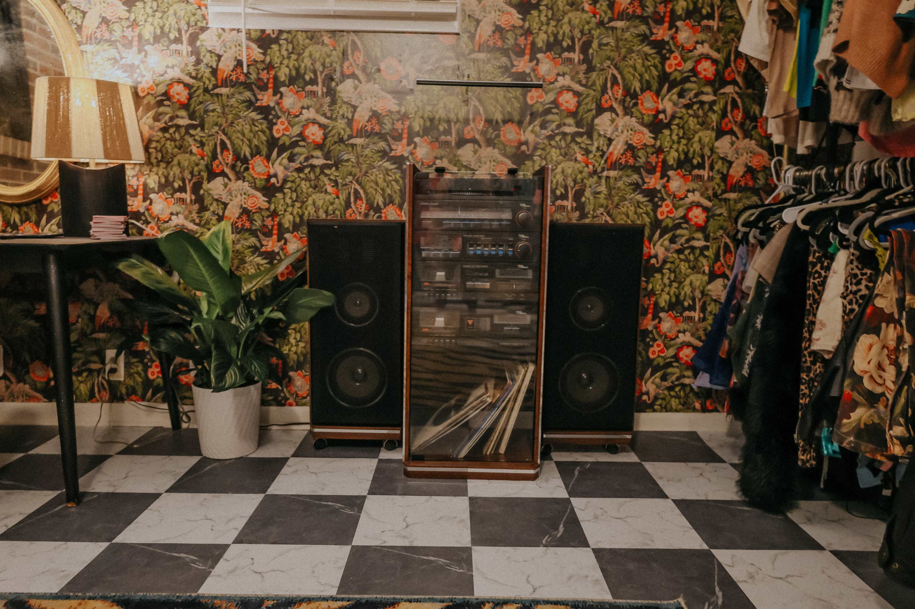 The image shows a wooden stereo system flanked by large speakers, against a floral wallpaper background, with a plant and a lamp nearby, all within a room featuring checkered flooring.