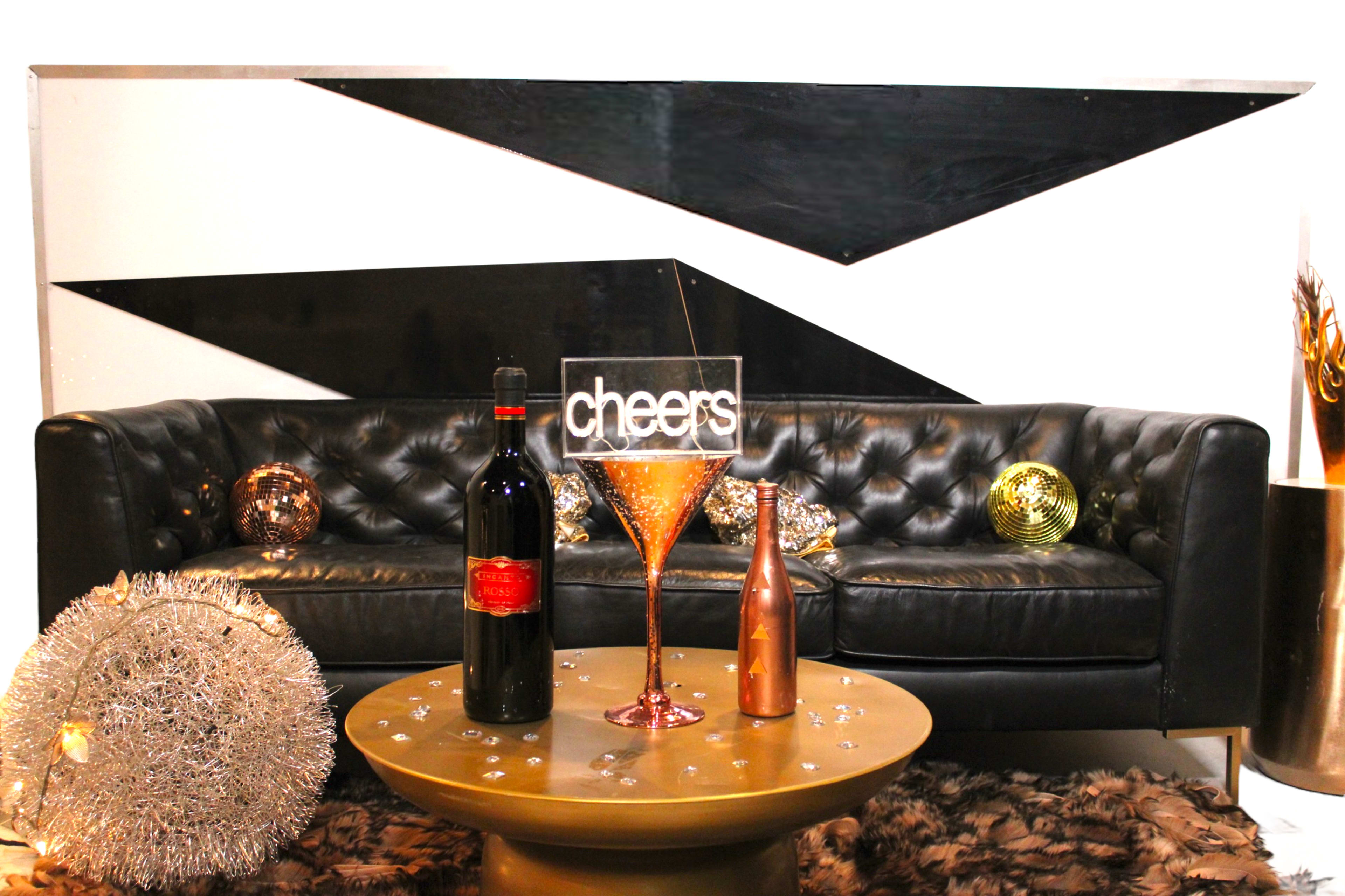 A black tufted sofa is positioned against a geometric black and white wall, with a bottle of wine, a large cocktail glass, and a small metallic bottle displayed on a round gold table.