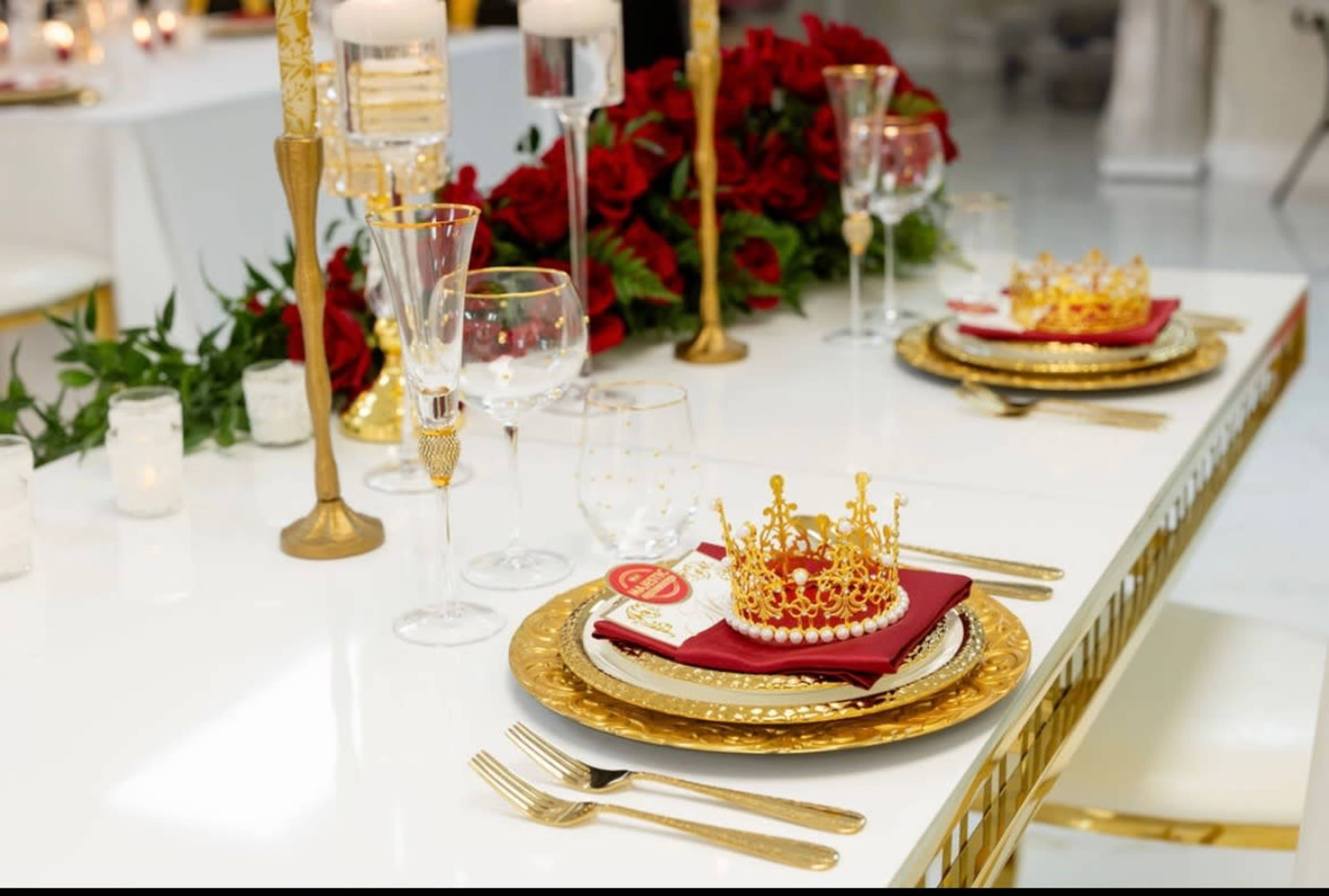The table is elegantly set with gold-accented dinnerware, crystal glassware, and decorative crowns resting on folded napkins surrounded by red roses.