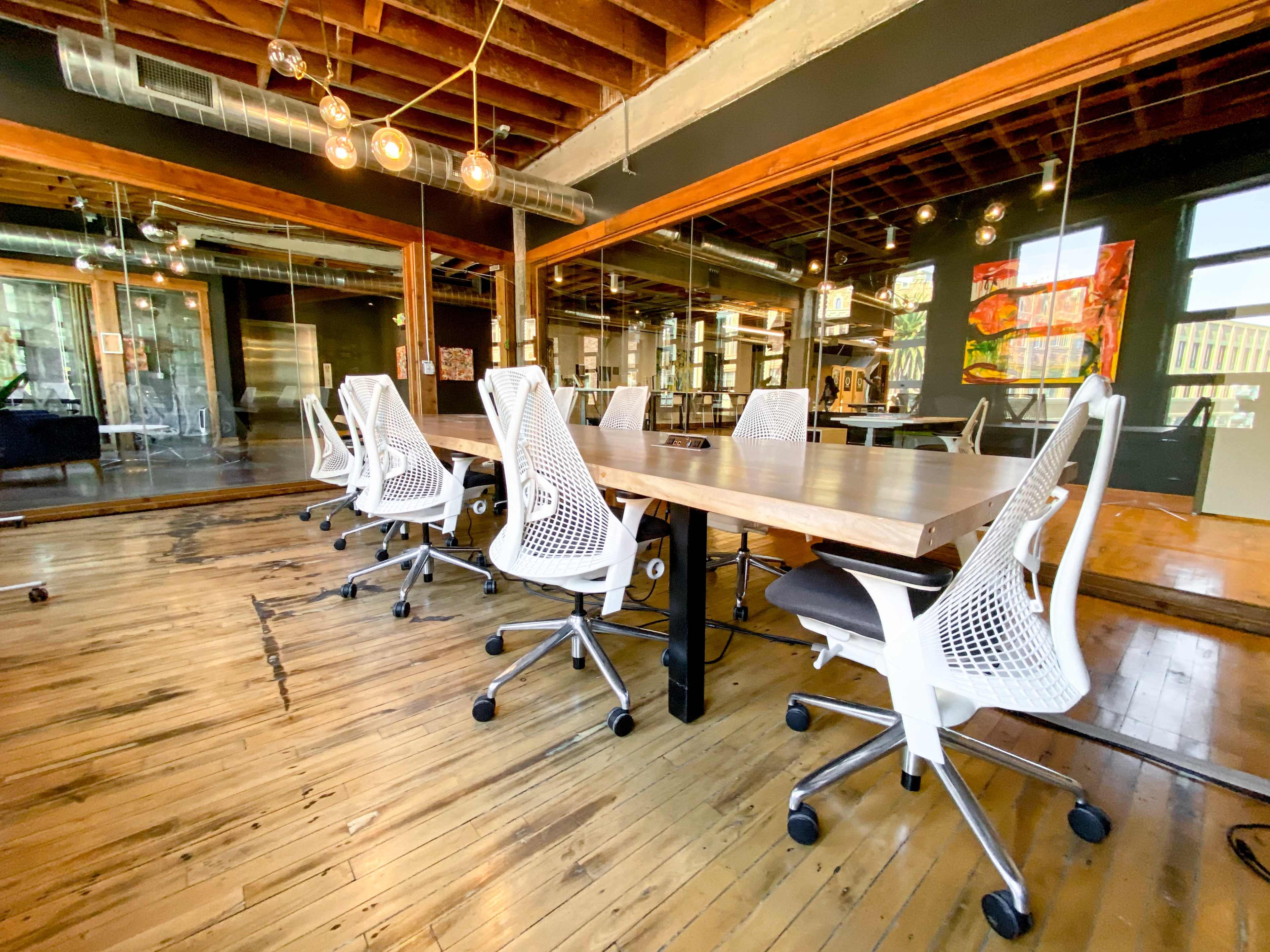 A conference room features a large wooden table surrounded by white office chairs, with exposed wooden beams and mirrors on the walls.