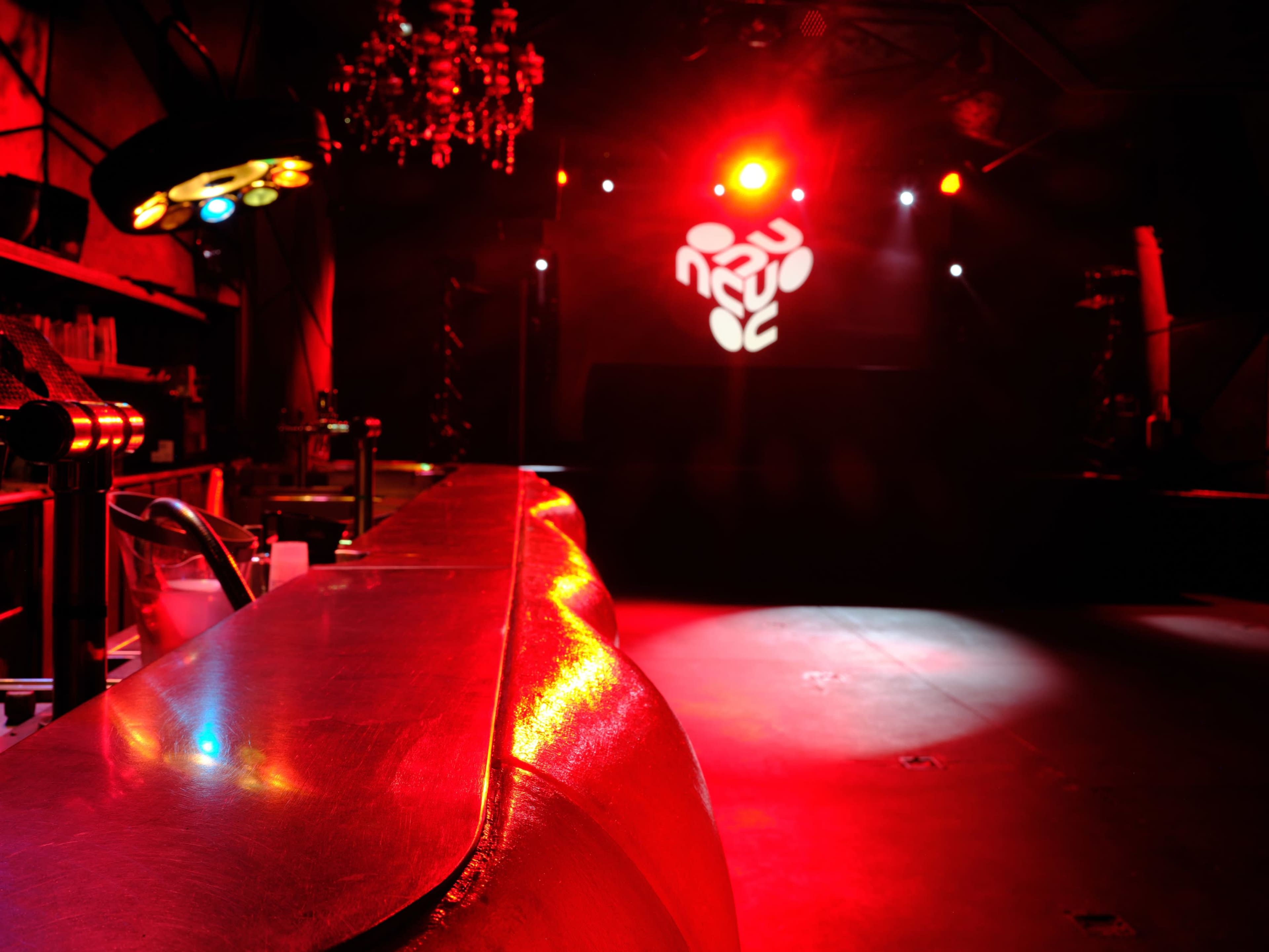 The image shows a dimly lit nightclub interior with a reflective bar surface illuminated by red lights and a logo projected on the wall.