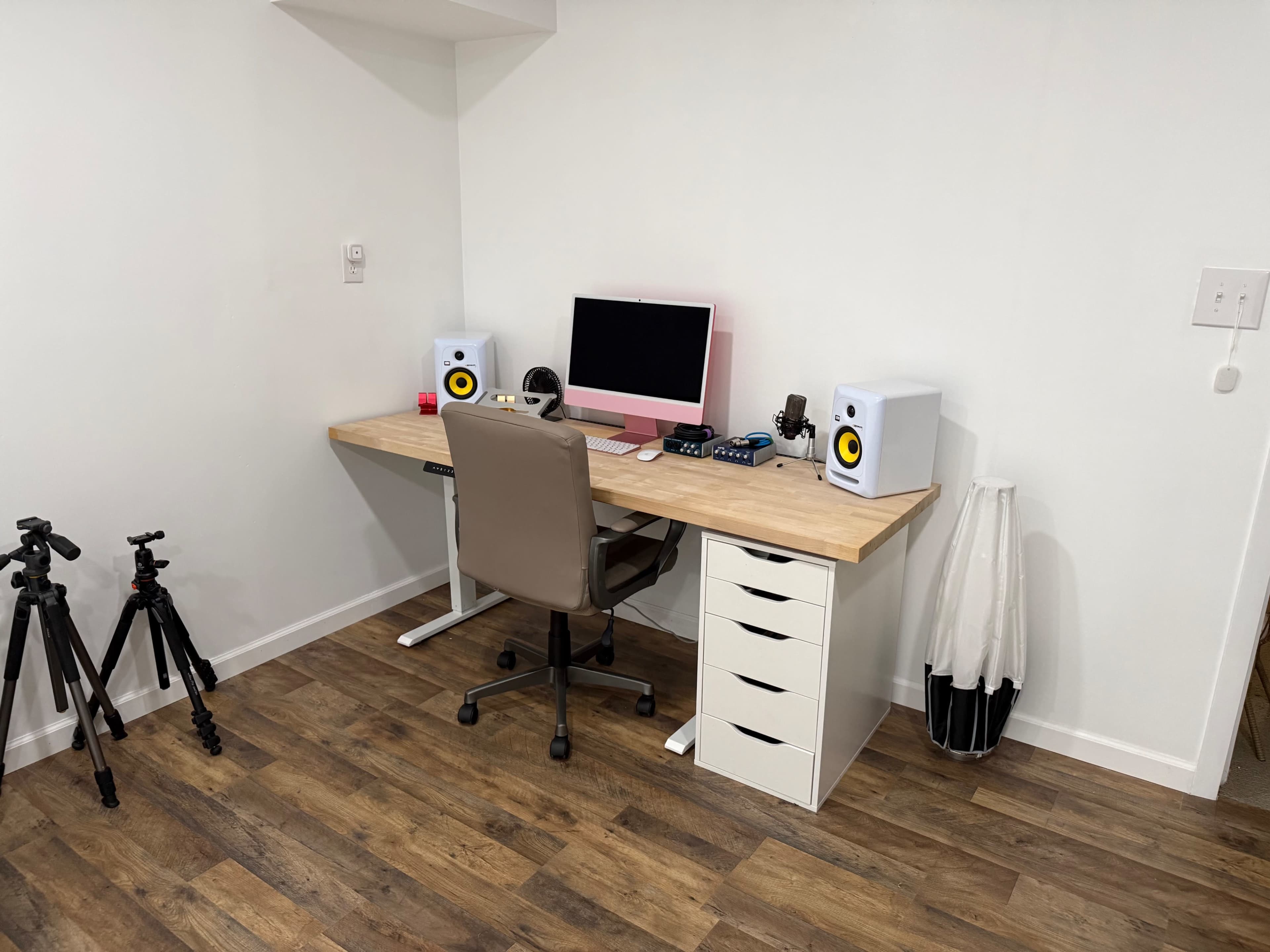The image shows a minimalistic home office setup featuring a wooden desk with a pink monitor, two audio speakers, a chair, and storage drawers, alongside a tripod and a white lamp.