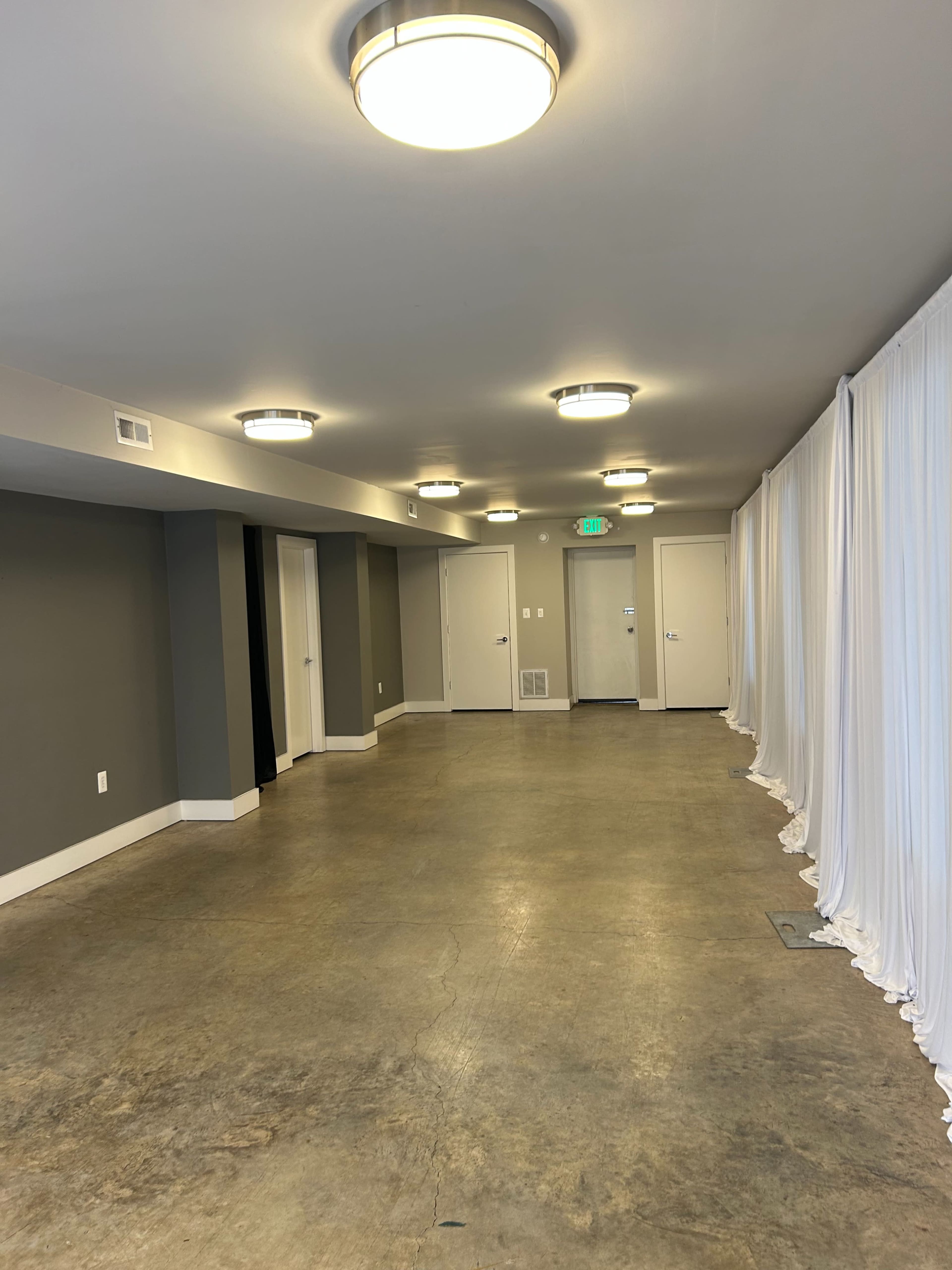 The image shows a long, empty hallway with gray walls, concrete flooring, and ceiling lights, leading to several doors and a white curtain on the right side.