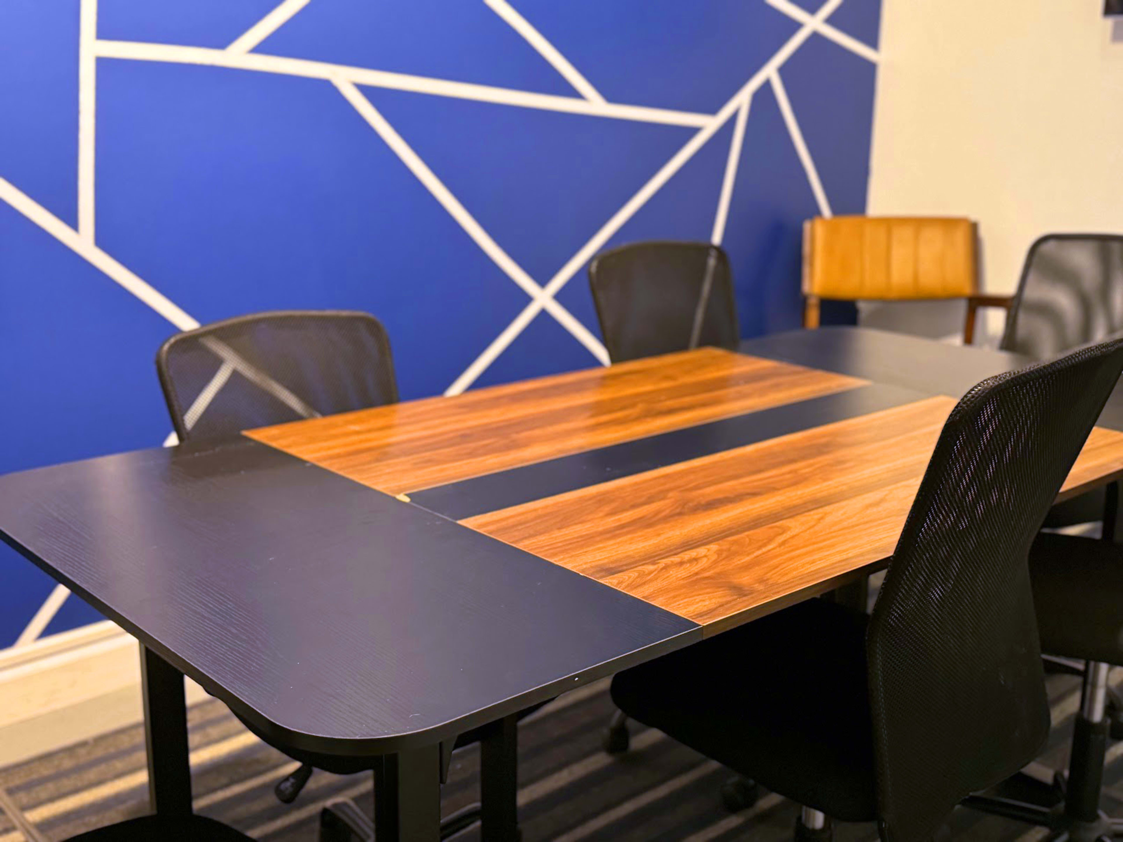 A conference room table with a wooden top and black edges is surrounded by four mesh chairs against a blue wall featuring white geometric patterns.