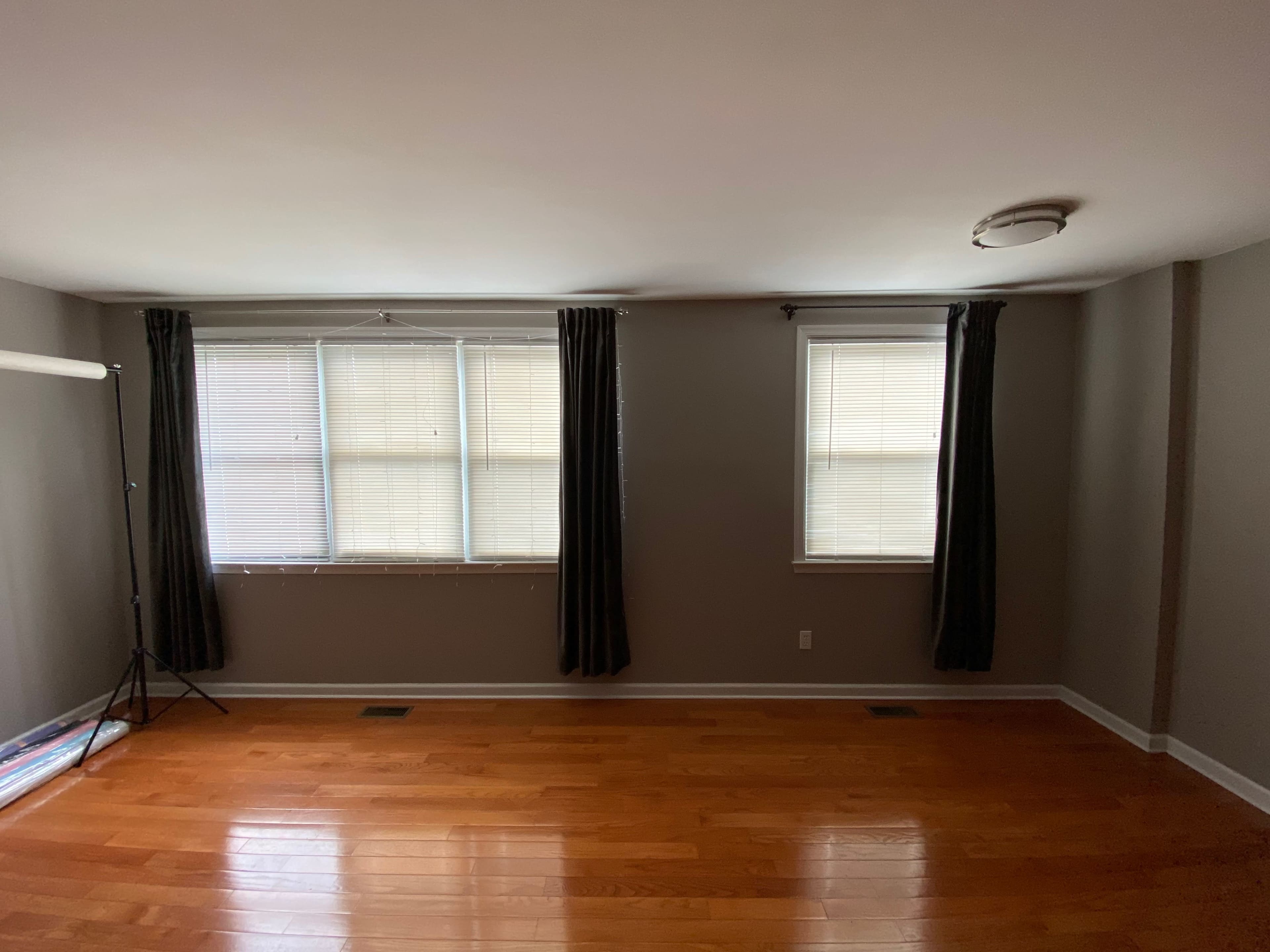 A sparsely furnished room with hardwood floors, two windows covered by closed blinds, and dark curtains on either side.