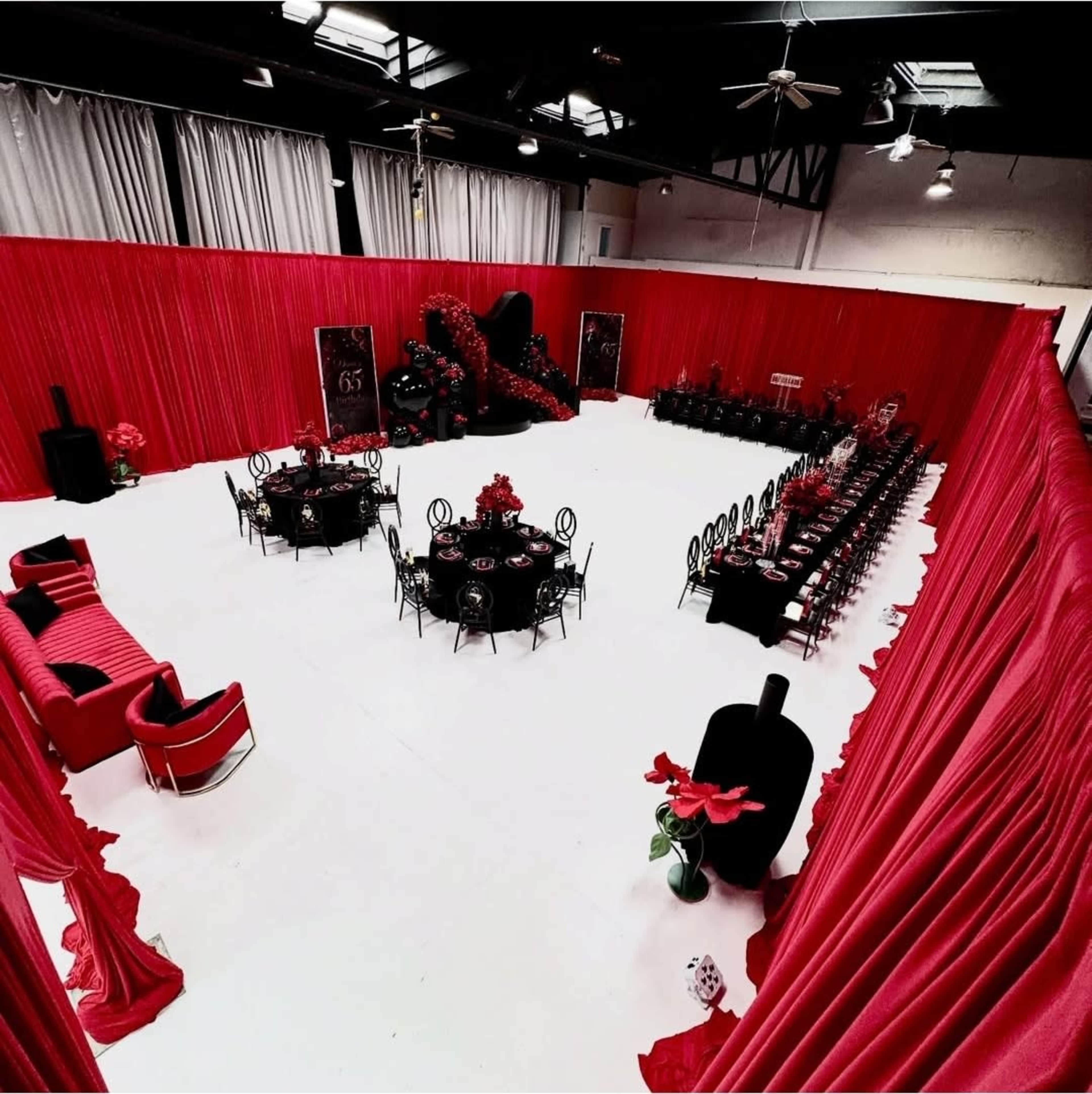 The image shows a large event space decorated with red curtains, multiple dining tables set with black and red tableware, and elegant seating arrangements.
