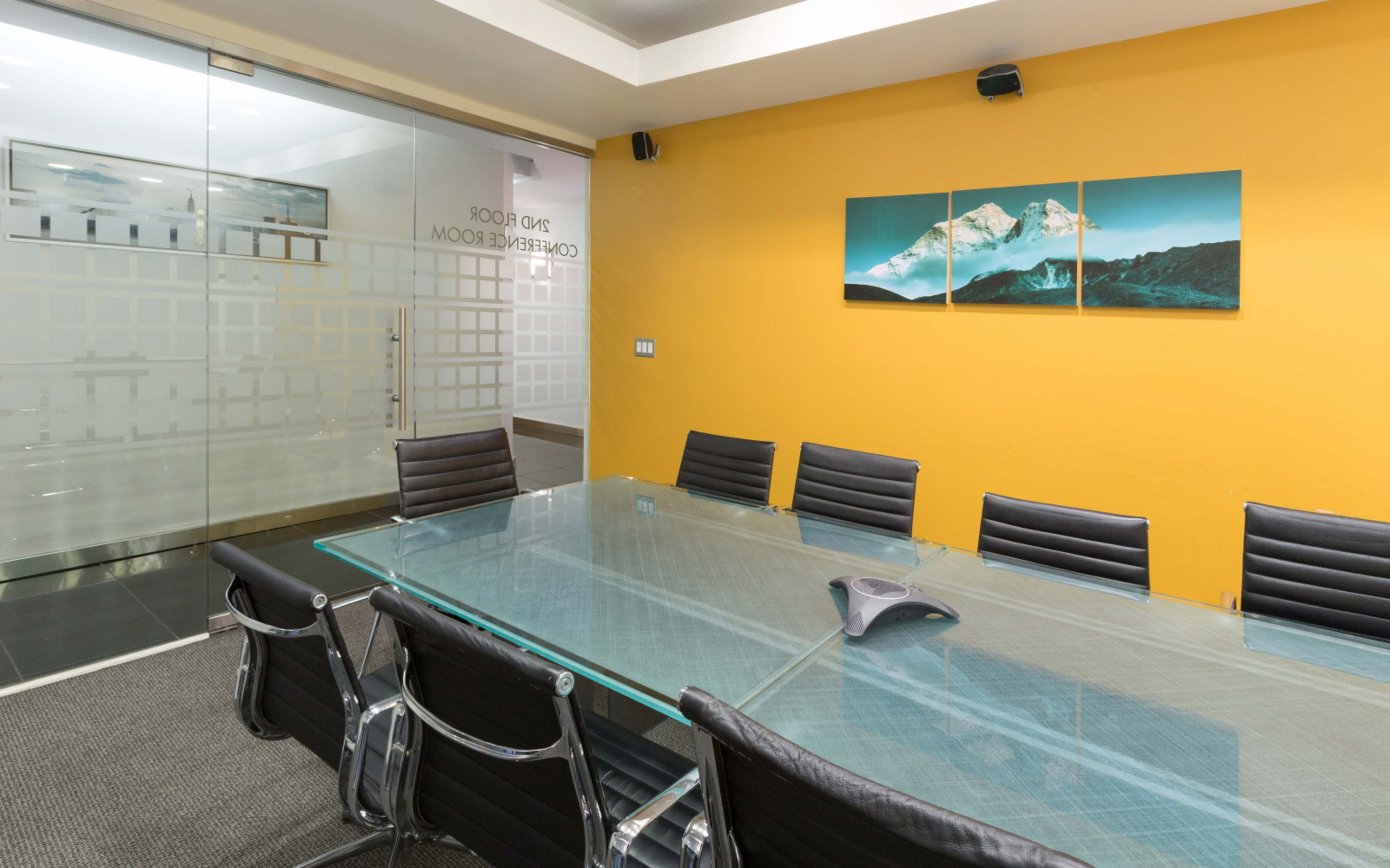 A glass conference table surrounded by black leather chairs is set against a bright orange wall, with a mountain landscape artwork displayed above the table.