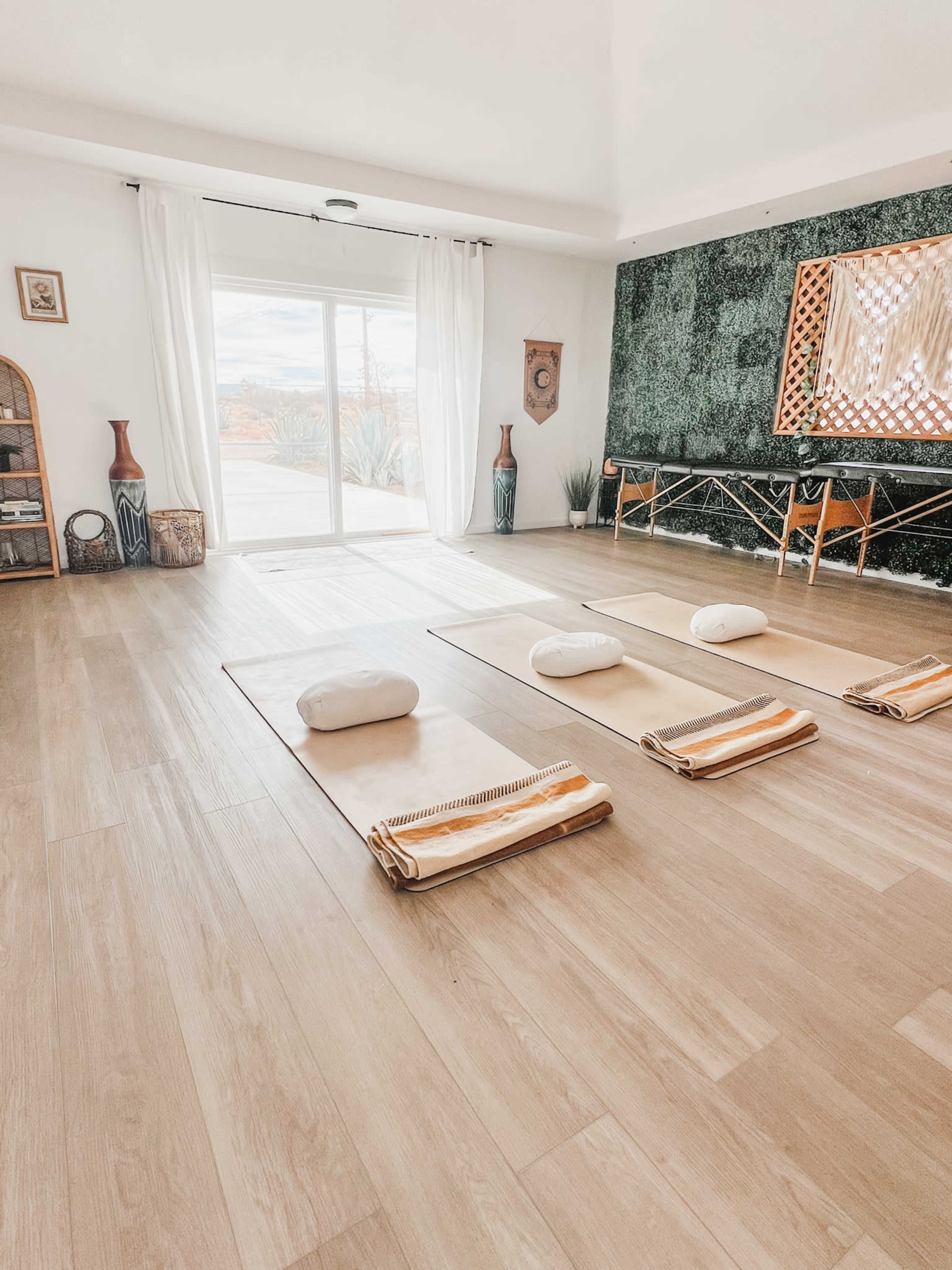 The image depicts a serene interior space designed for relaxation, featuring mats with cushions arranged neatly on a wooden floor, large windows allowing natural light, and decorative elements on the walls.