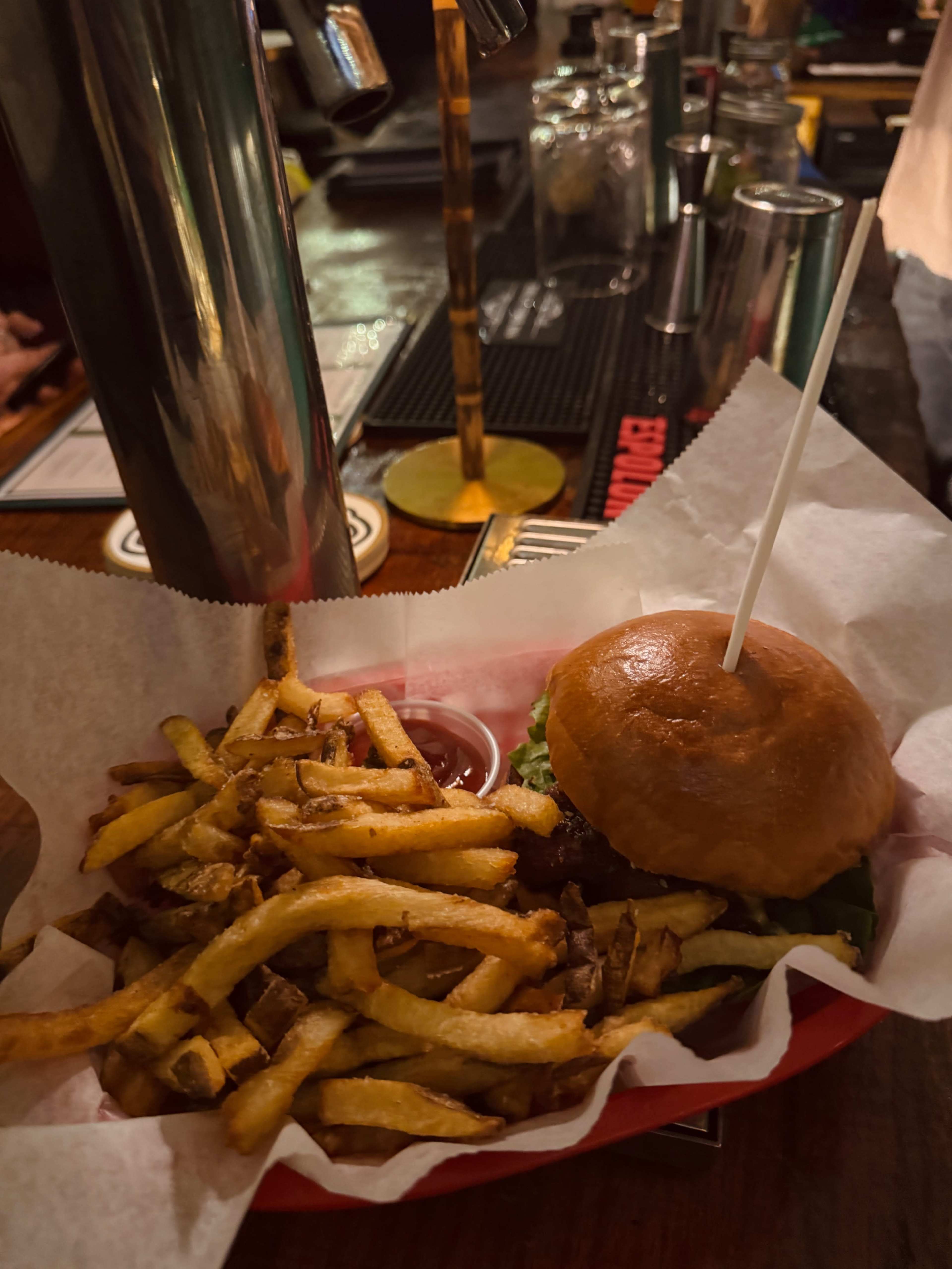 A hamburger with lettuce is served in a red basket alongside a portion of golden fries and a small cup of dipping sauce.