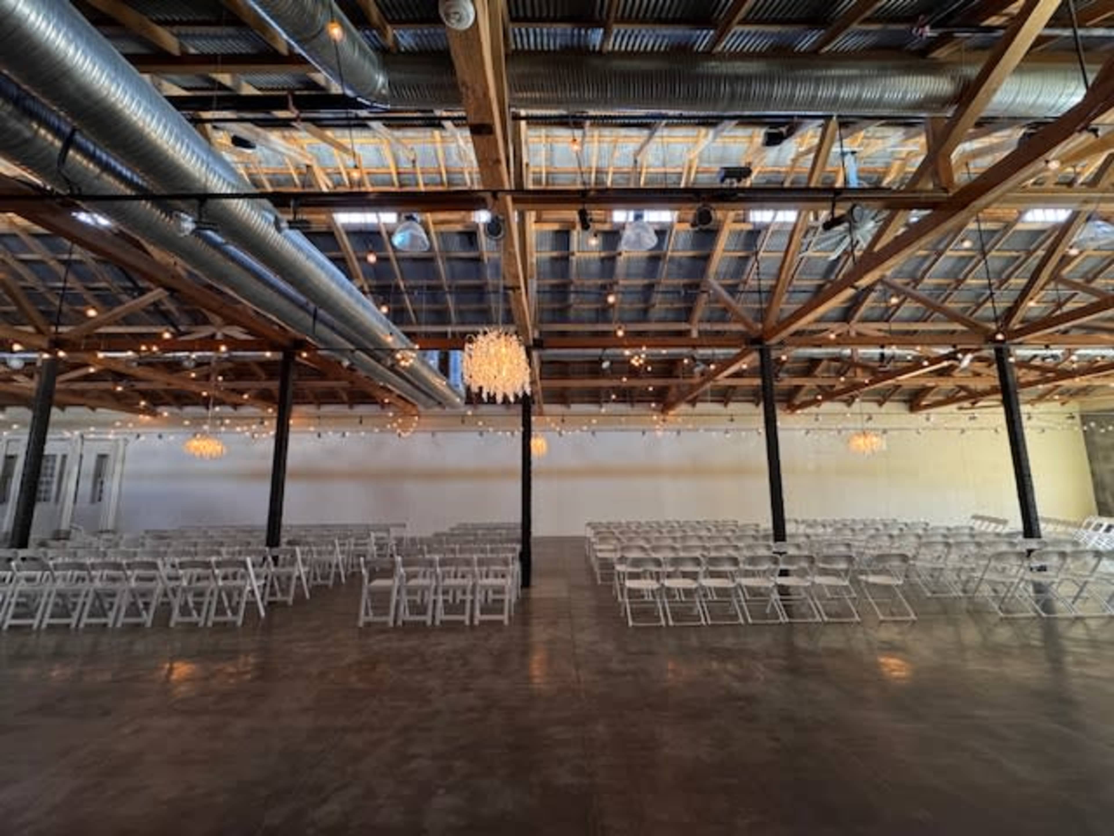 The image shows a spacious event venue with rows of white folding chairs arranged for seating, illuminated by overhead lights and decorated with a chandelier.