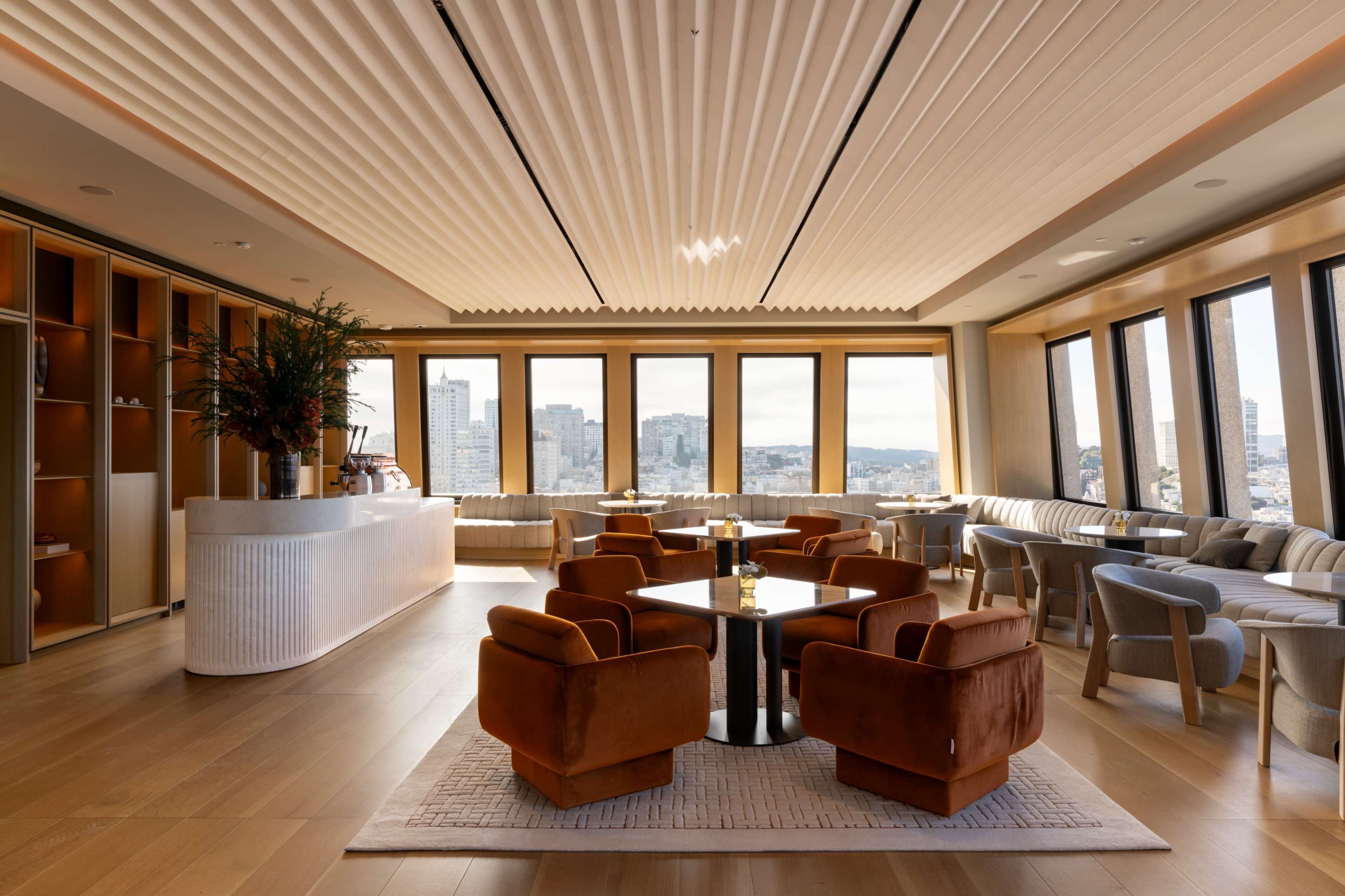 The image shows a stylish lounge area with modern furniture, large windows offering a city view, and a reception counter.