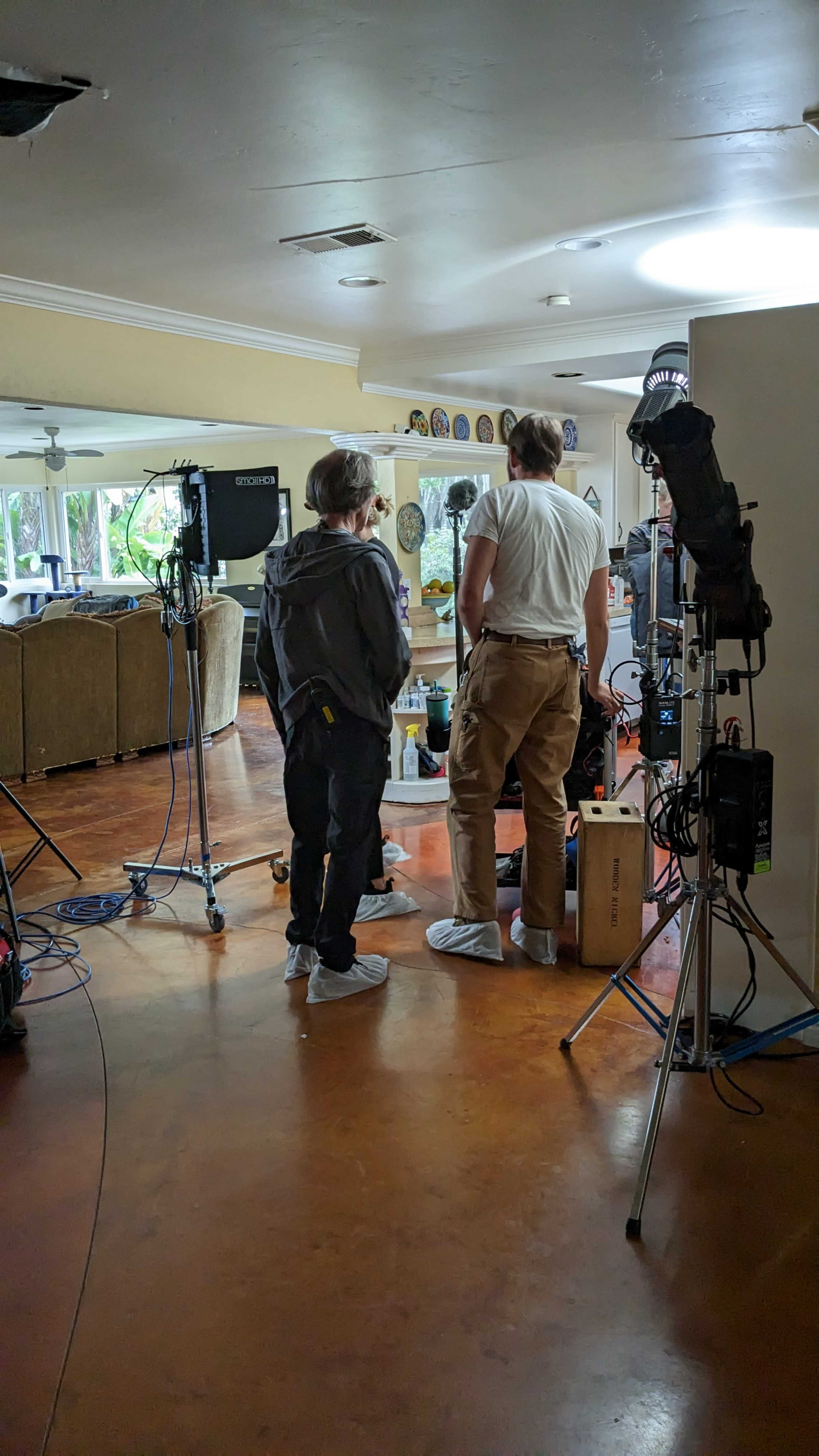 Two crew members prepare equipment in a living room set for a film shoot, while cameras and lighting gear are arranged nearby.