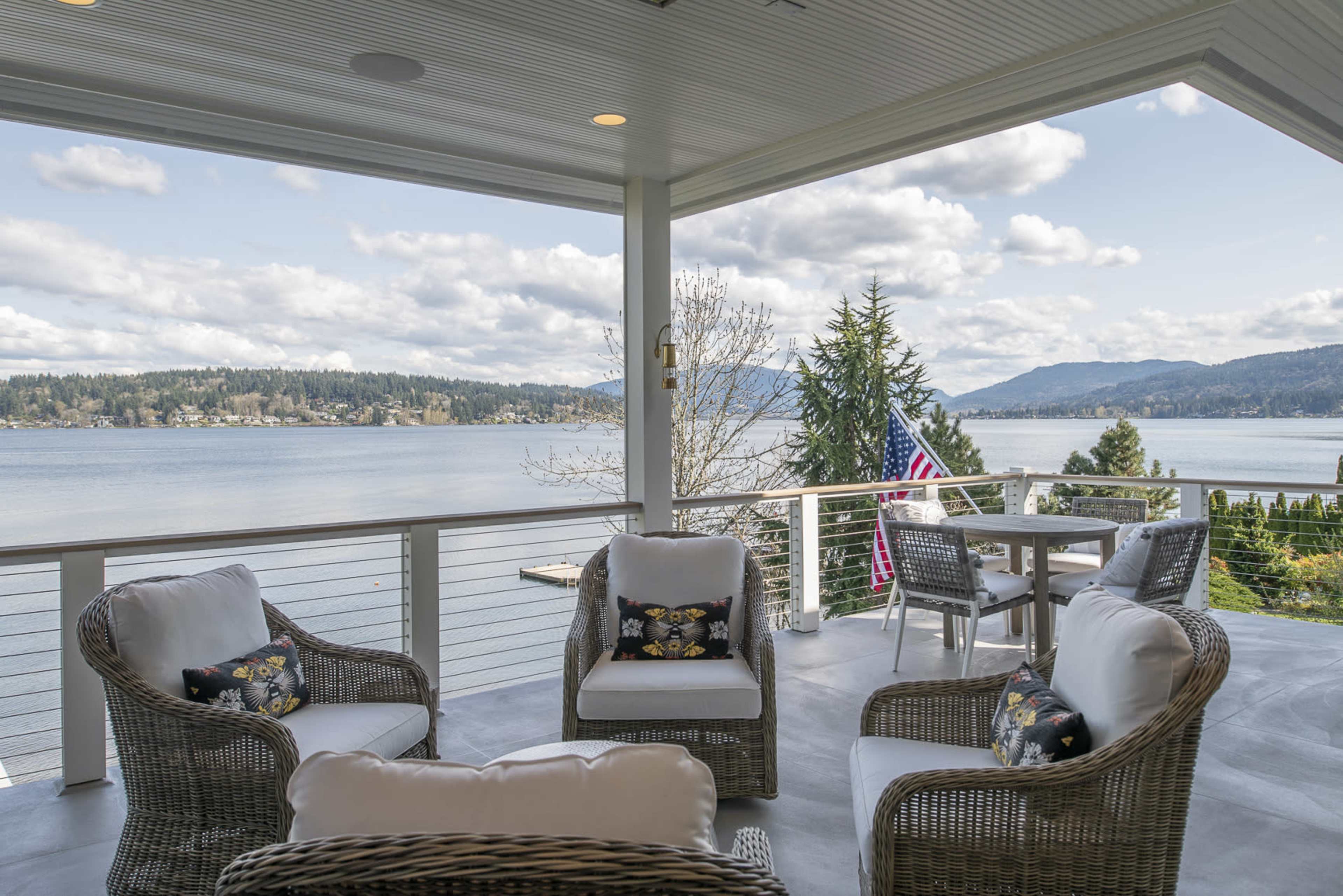 A covered patio with seating and a dining area overlooks a lake and mountains in the background.