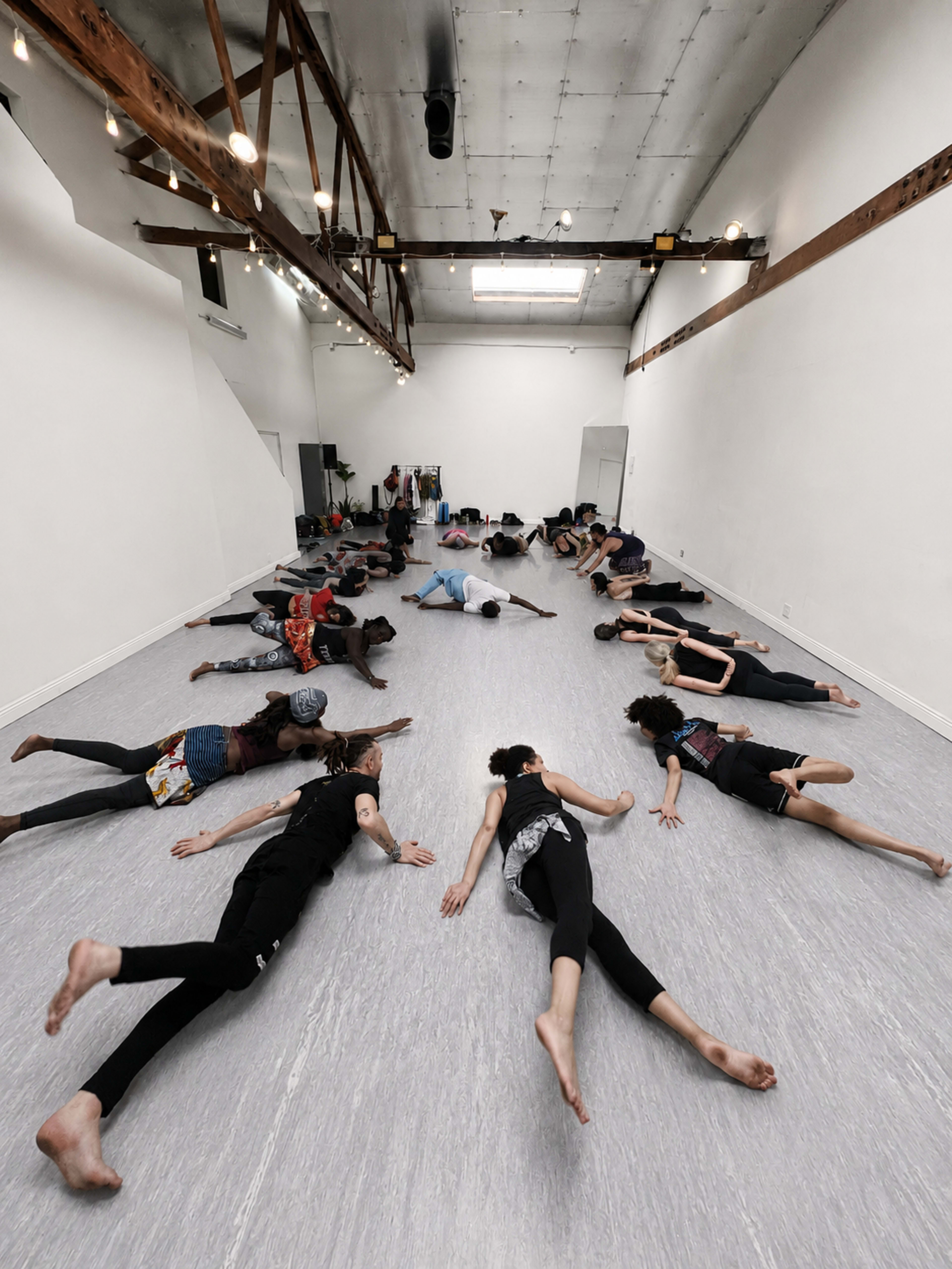 A group of people is lying on the floor in a large, open dance studio with a high ceiling and exposed wooden beams.