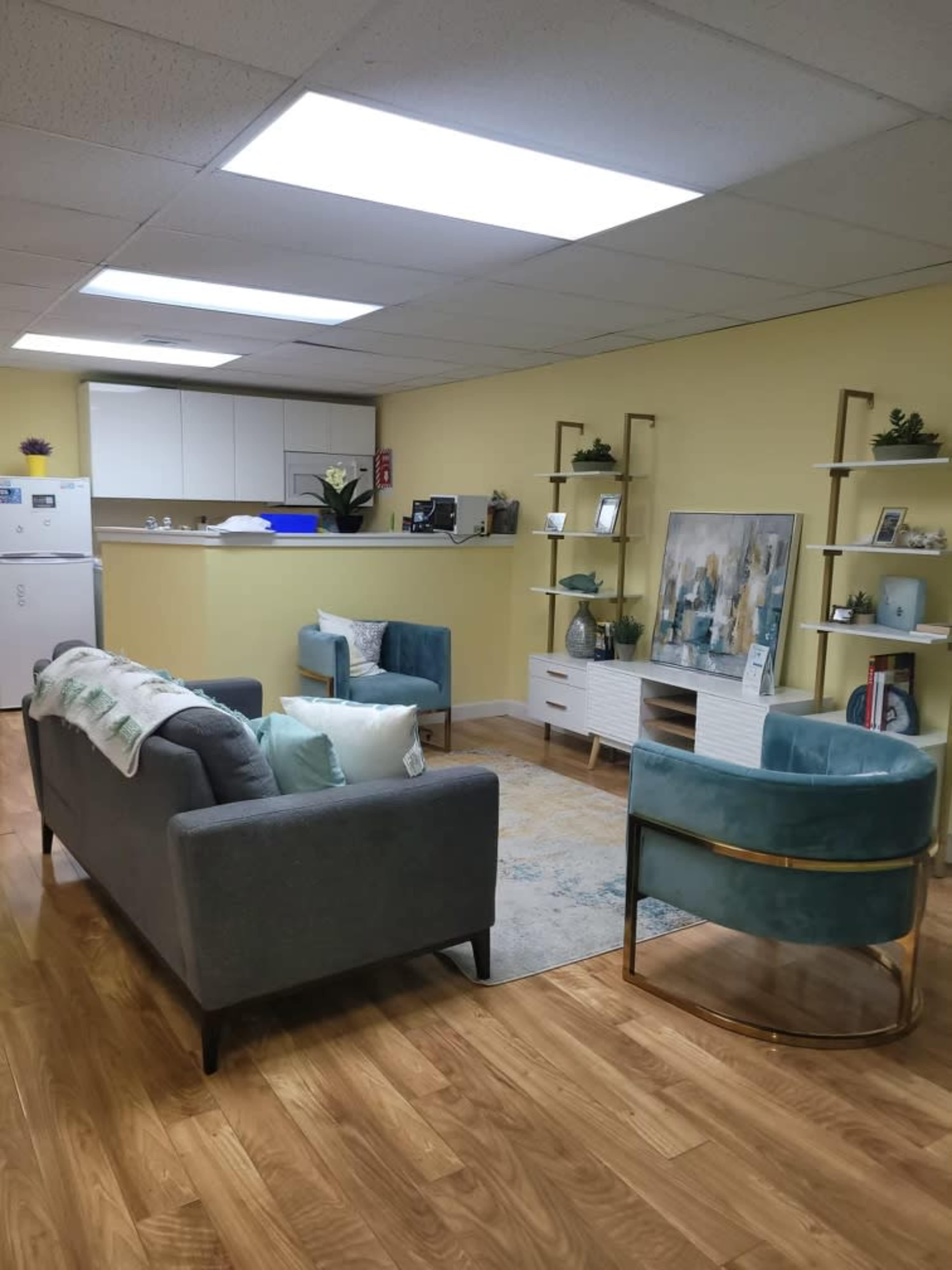 A modern office space with a seating area featuring two armchairs and a sofa, a coffee table, and a kitchenette in the background.