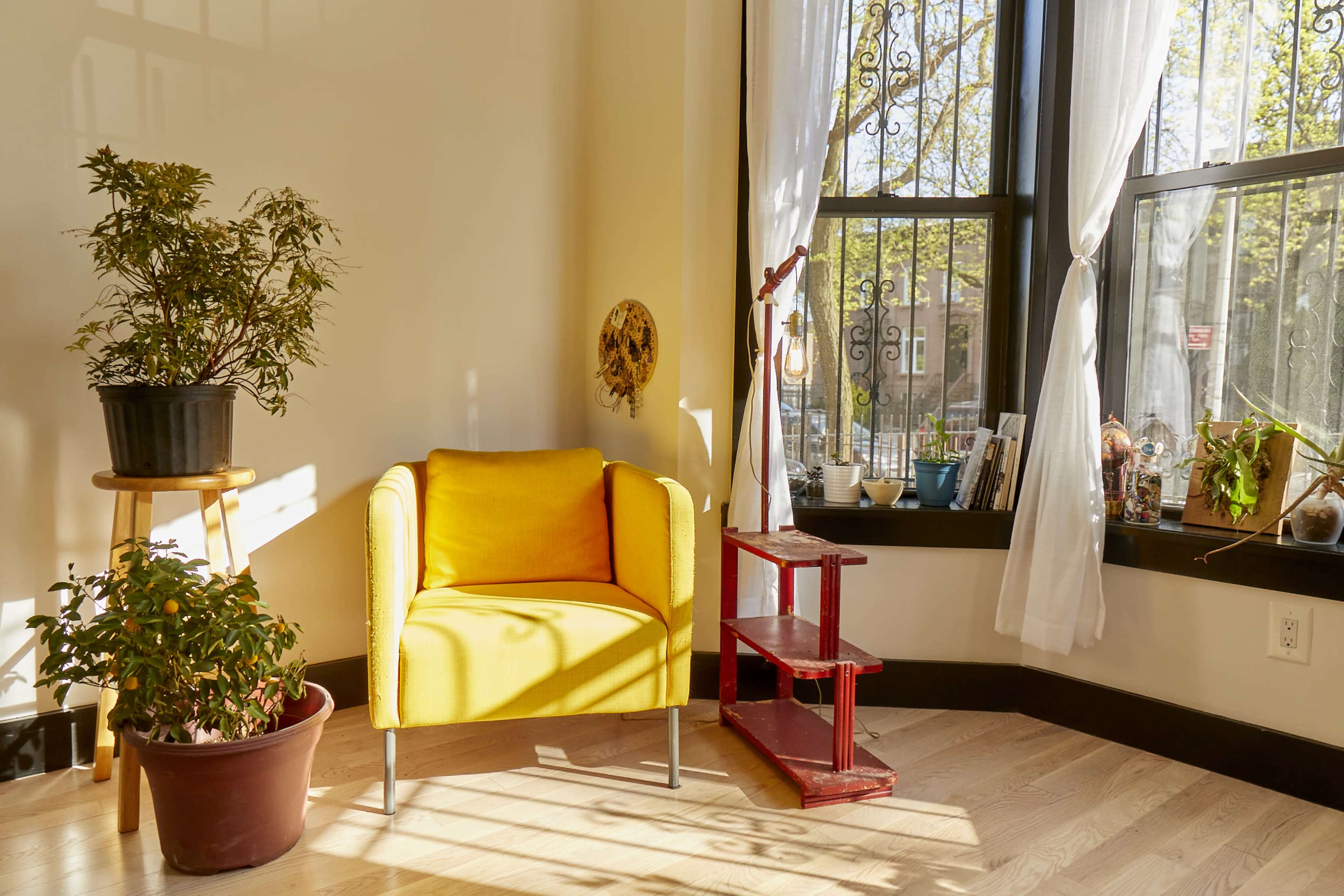A yellow armchair is positioned beside a window with flowing white curtains, next to a potted plant and a red wooden shelf displaying various items.
