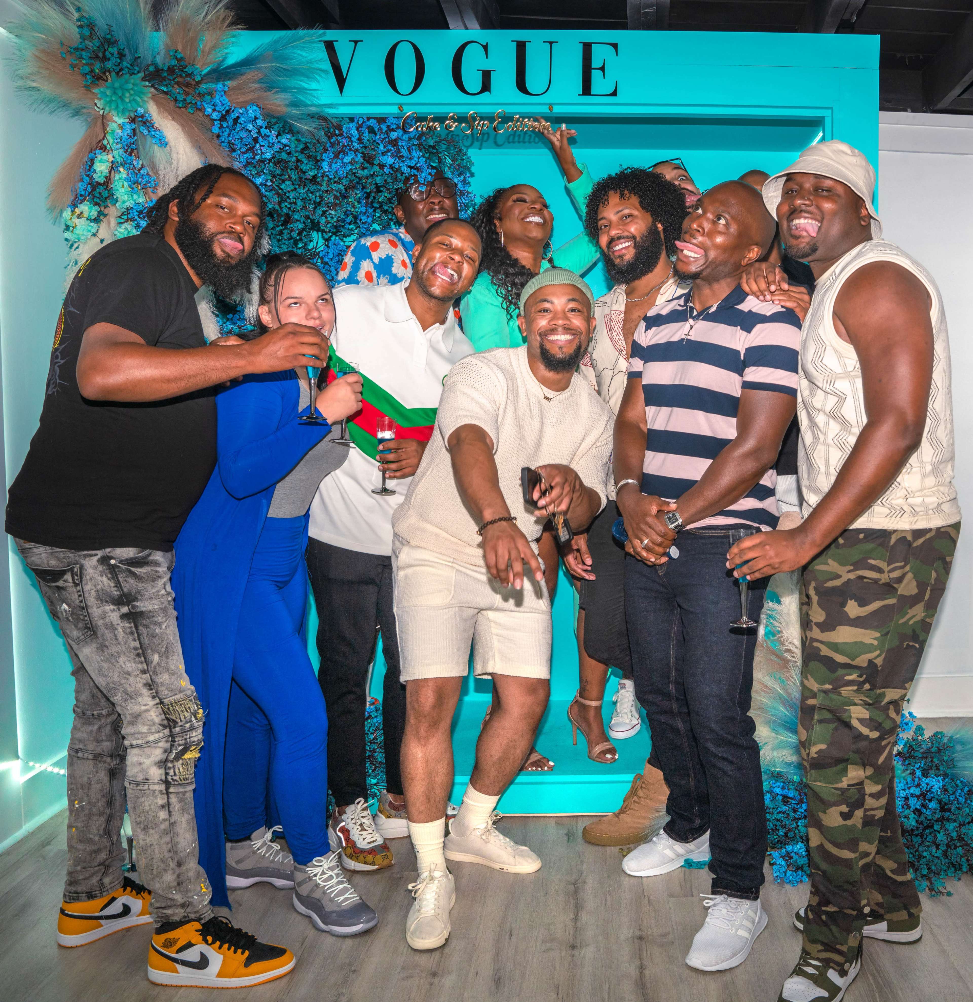 A group of nine people pose together in front of a VOGUE sign, smiling and enjoying their time at an event.