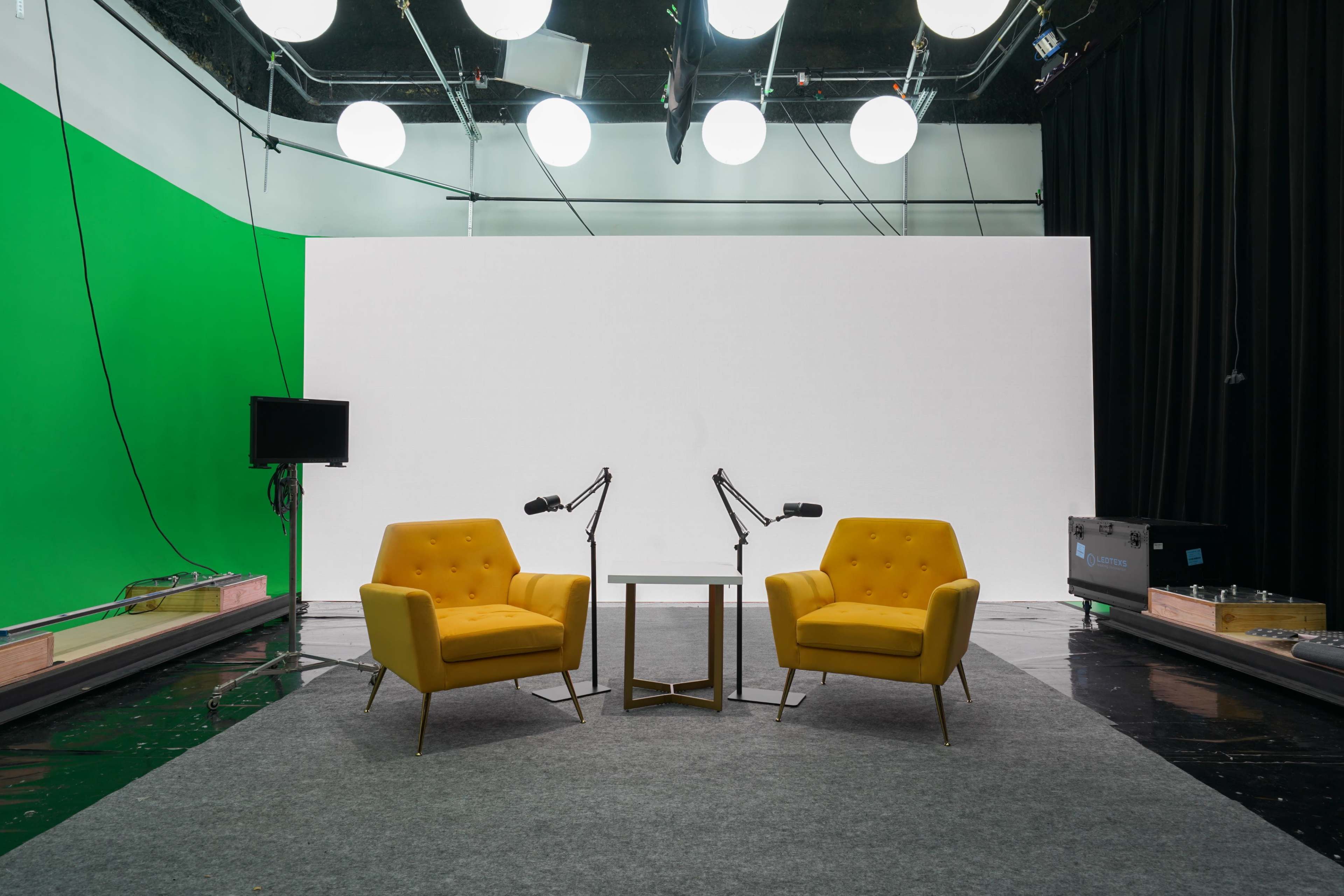 The image shows a studio setup with two yellow armchairs facing a small table, surrounded by a grey carpet and illuminated by large overhead lights against a green and white backdrop.