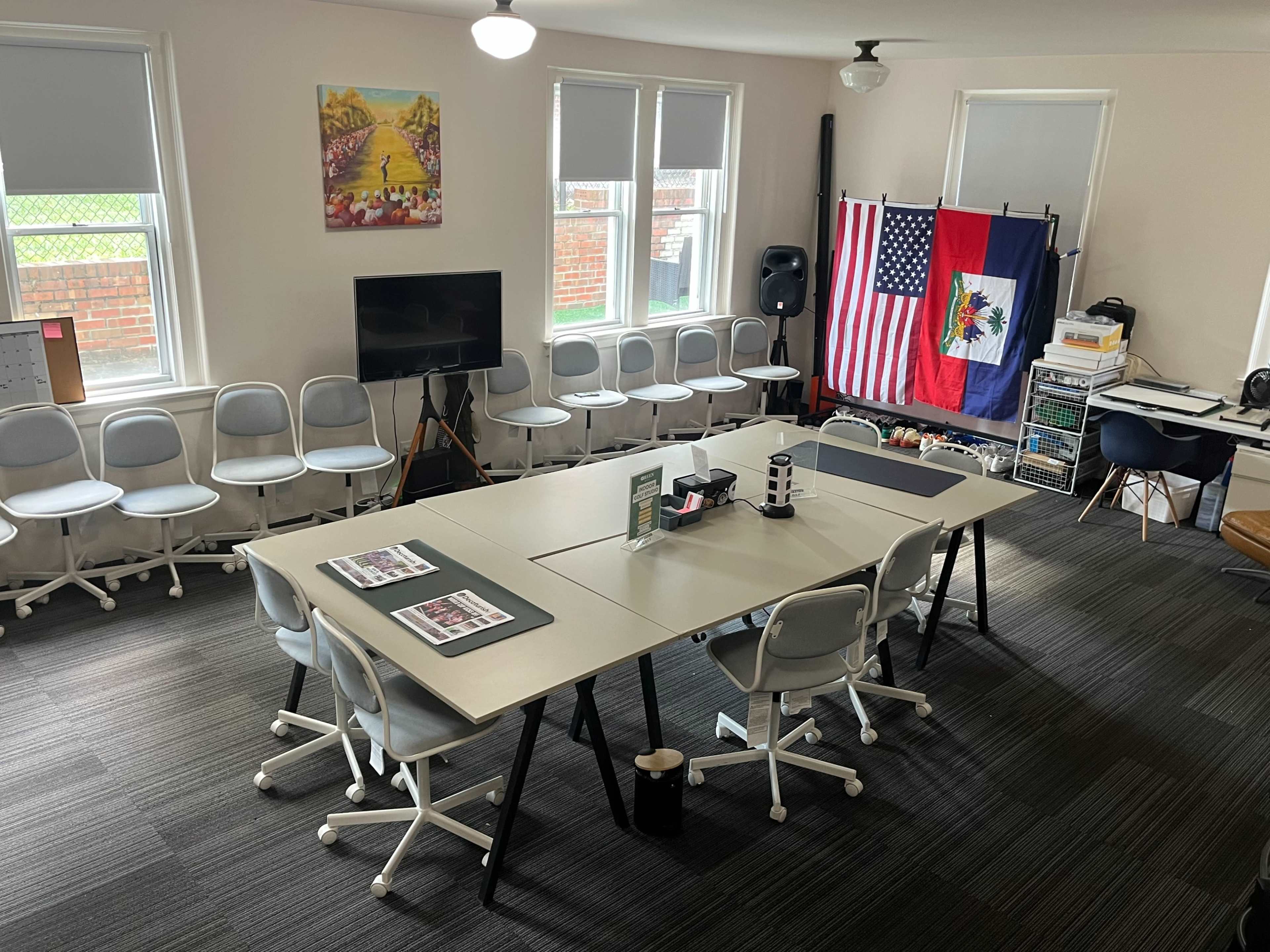 The image shows a spacious meeting room with multiple tables arranged in a U-shape, chairs around them, a television, and flags displayed on a wall.