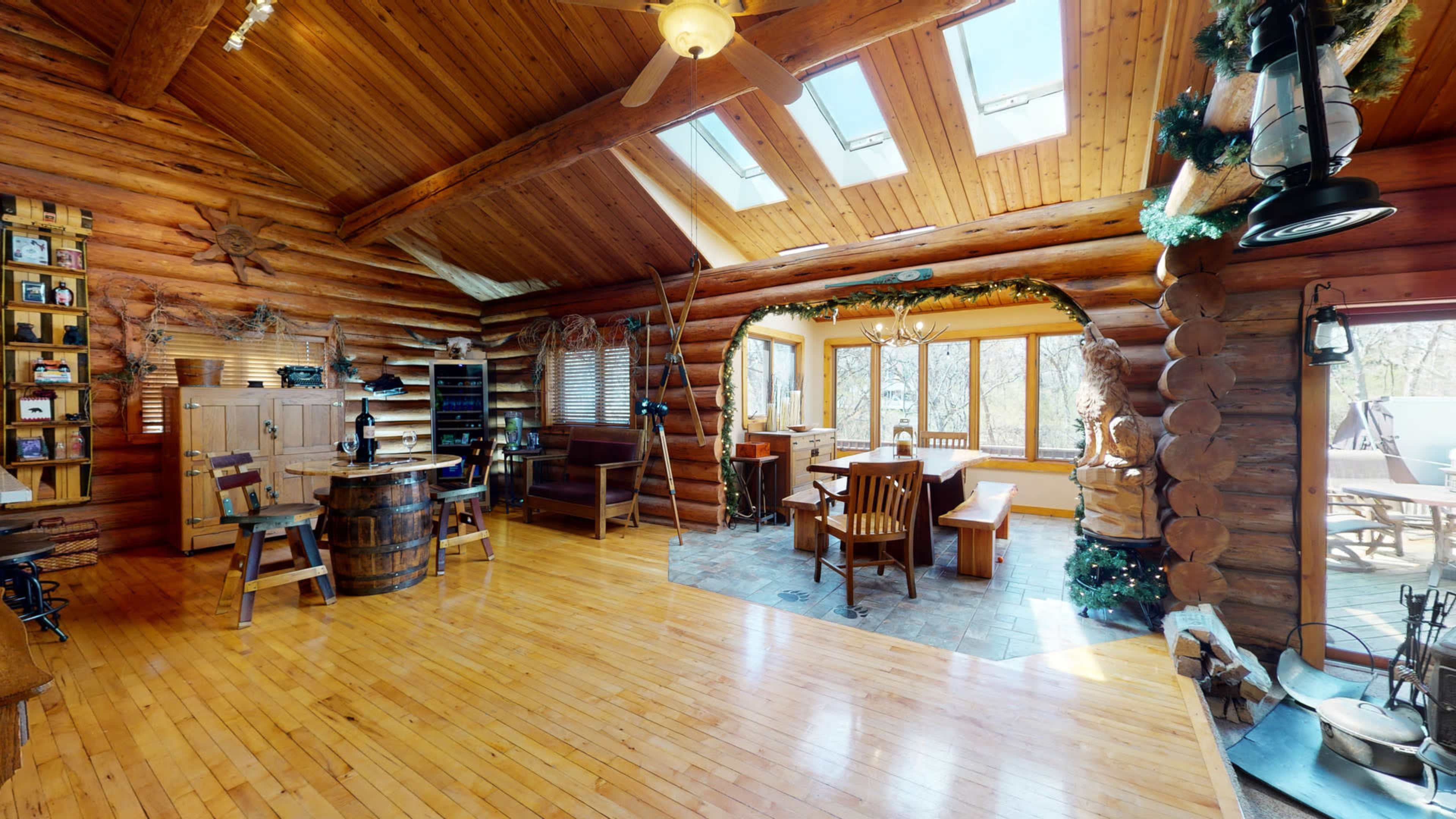 A spacious log cabin interior with wooden beams, large windows, a dining area, and a rustic decor theme.