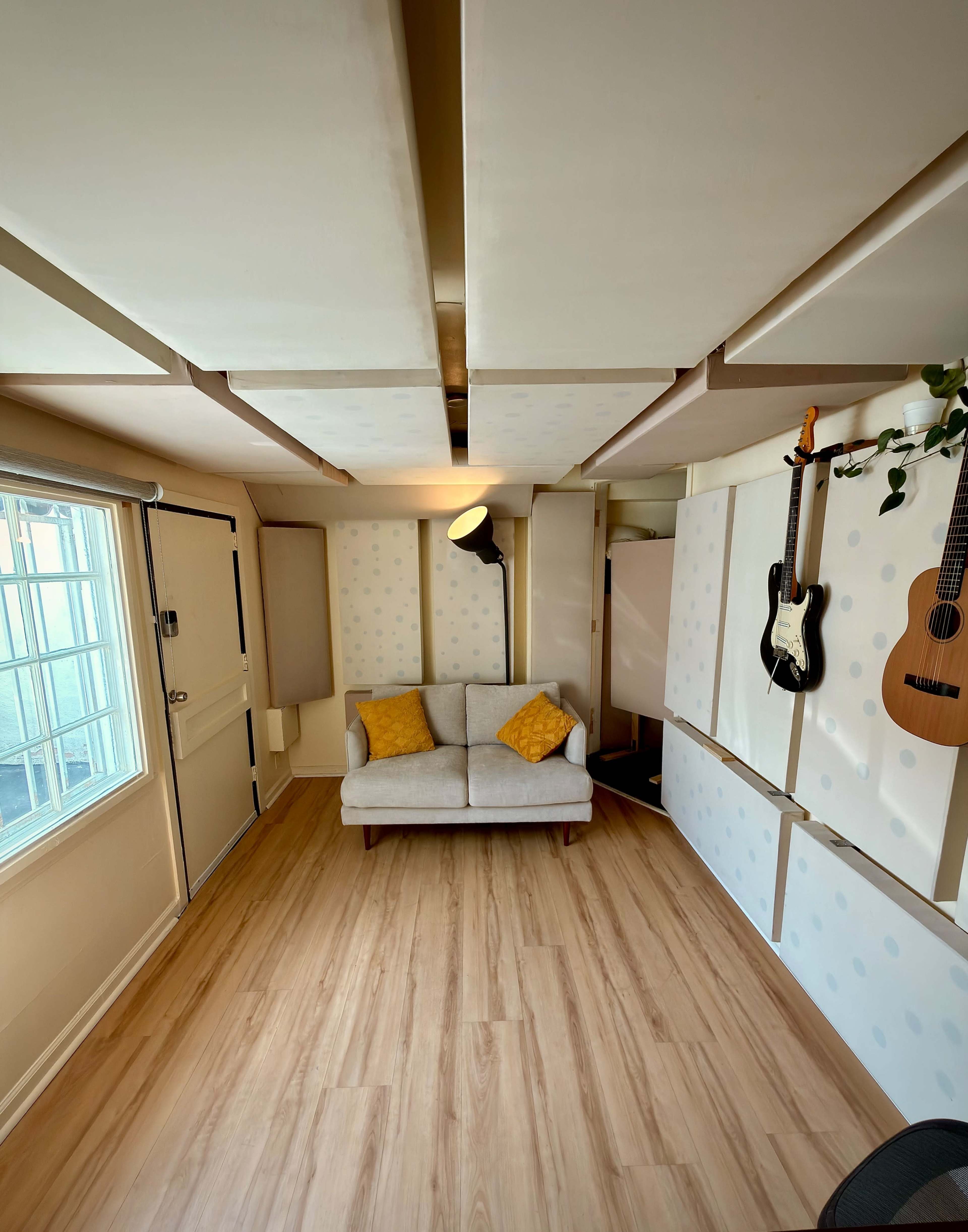 The image shows a small room with a light-colored couch, two decorative cushions, a lamp, an acoustic guitar hung on the wall, and a doorway leading outside.