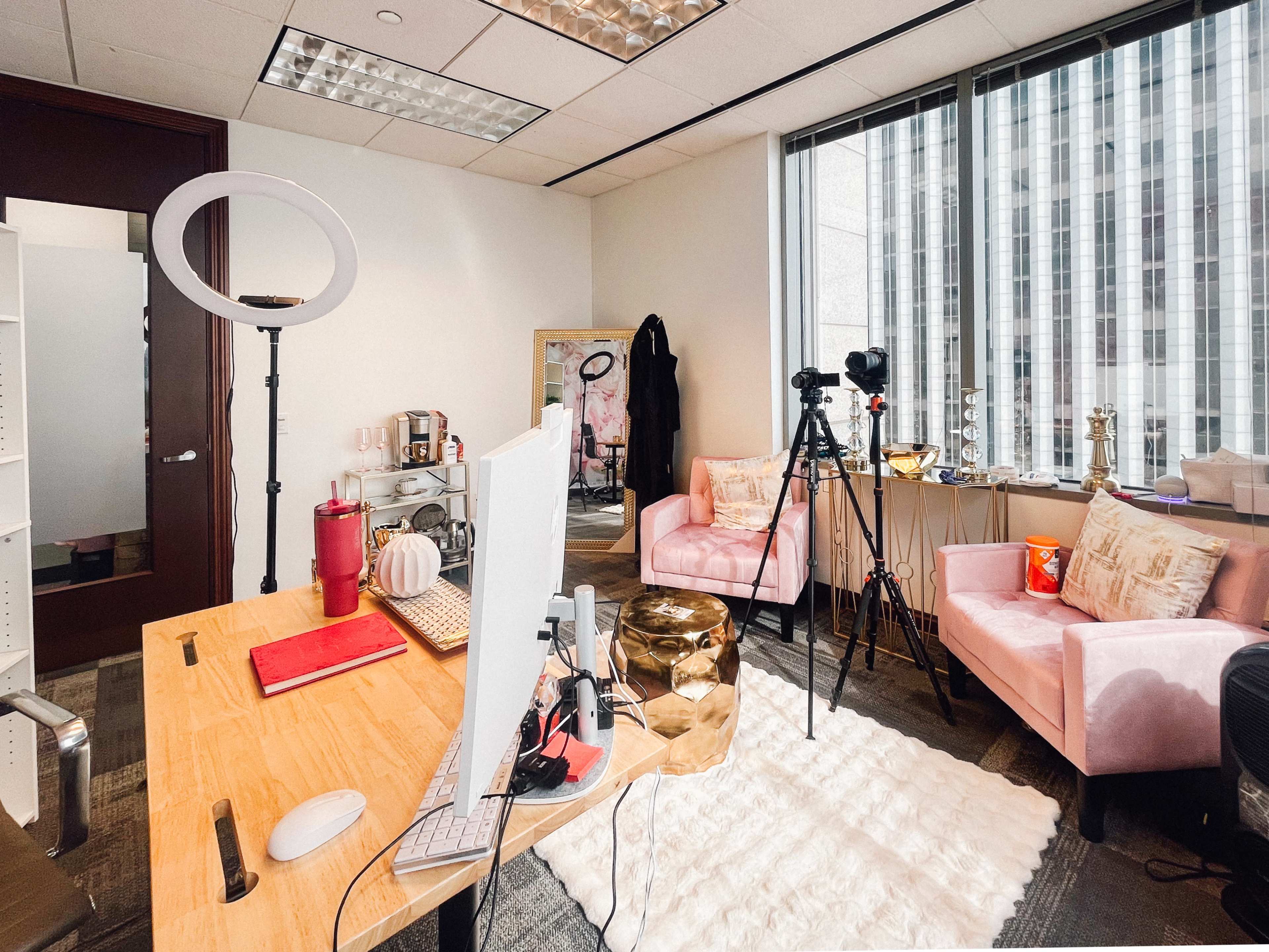 A modern office space features a wooden desk with a computer, two pink armchairs, cameras on tripods, and decorative items, all illuminated by natural light from large windows.