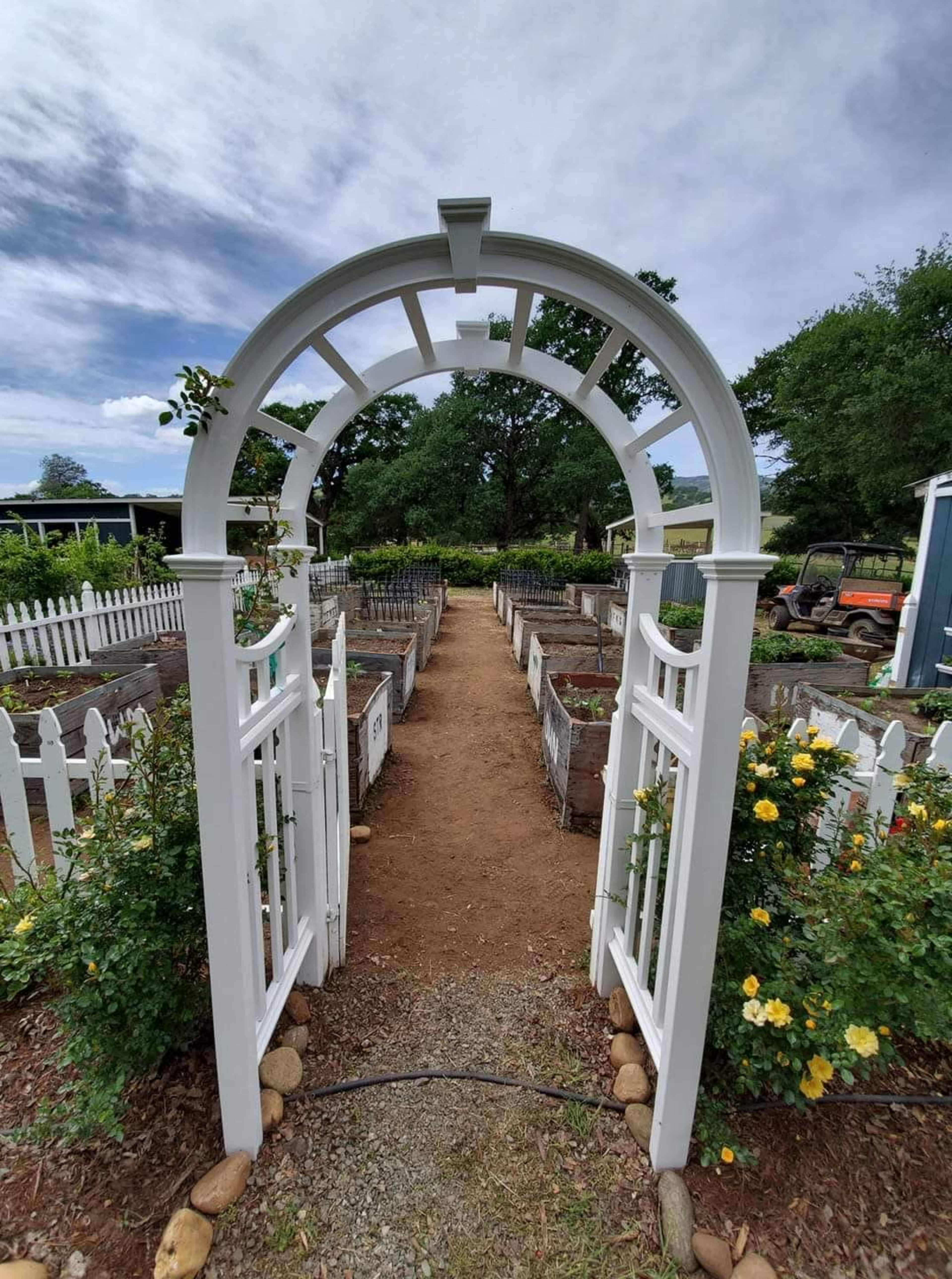 A white wooden arbor frames a dirt path leading through a garden filled with raised planting beds and surrounding greenery.
