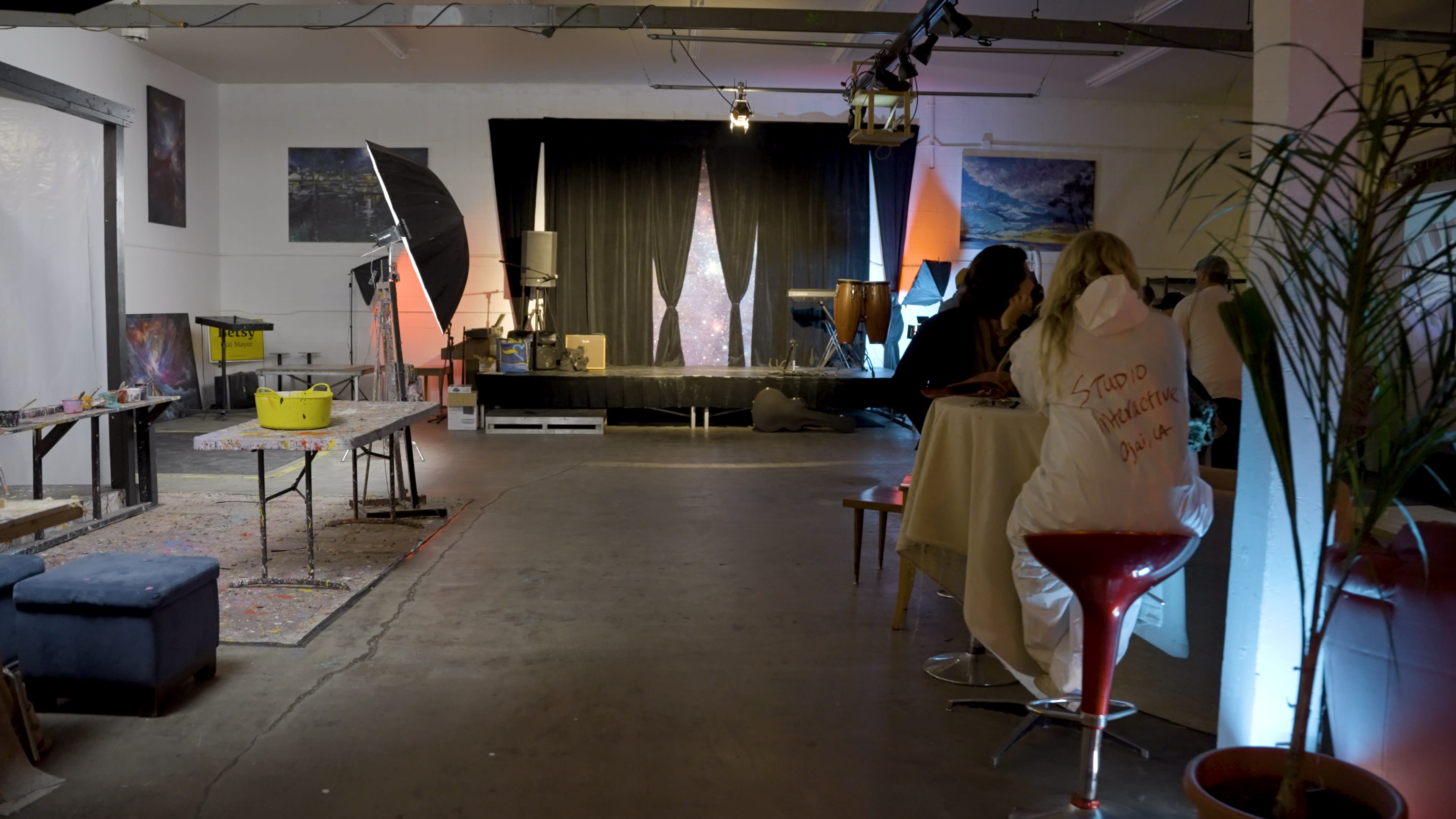 An art studio featuring a table, stools, and various art supplies, with a large black curtain at the back and paintings on the walls.