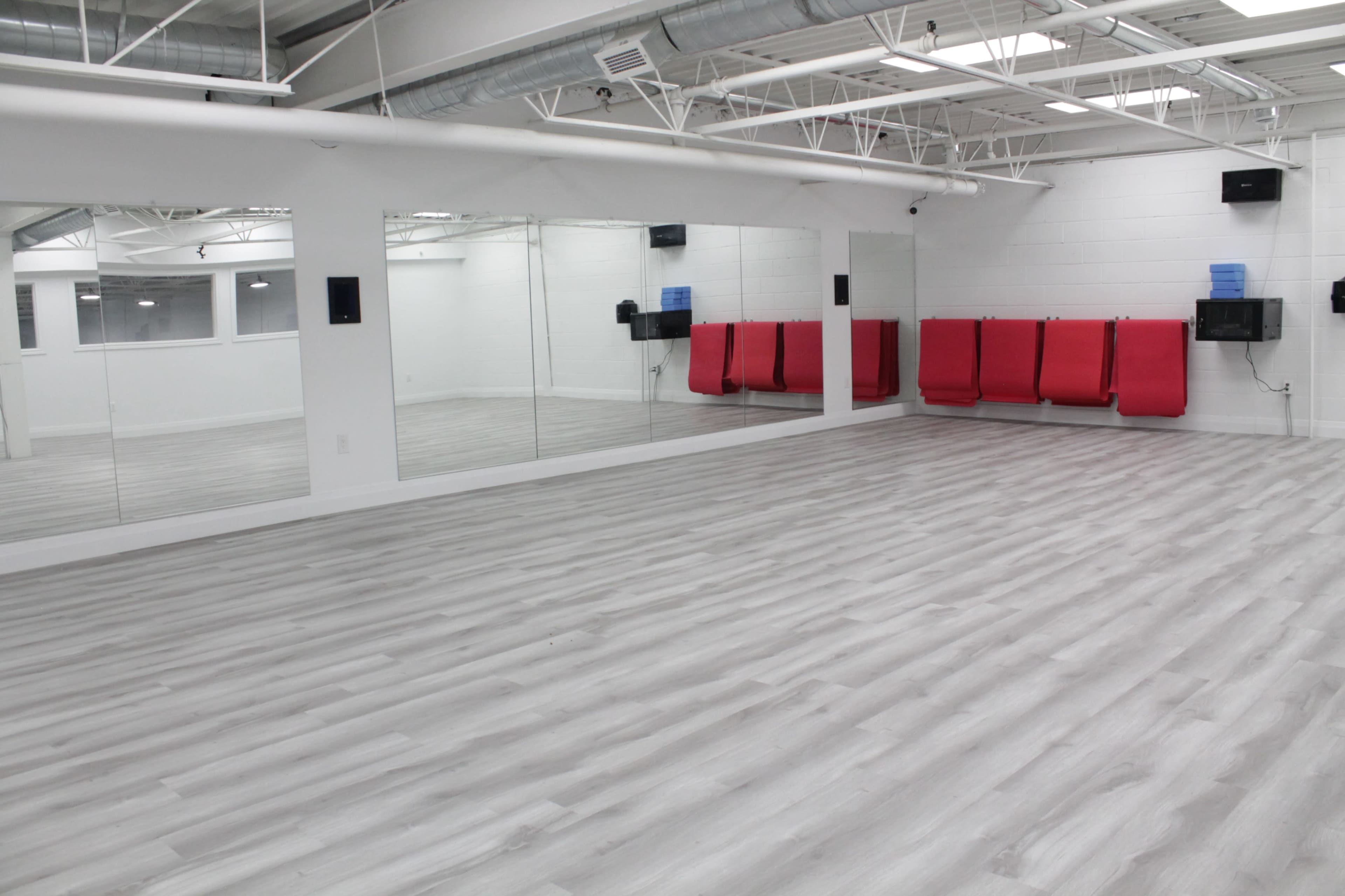 An empty, well-lit room with gray laminate flooring, white walls, a large mirrored wall, and red wall-mounted benches.