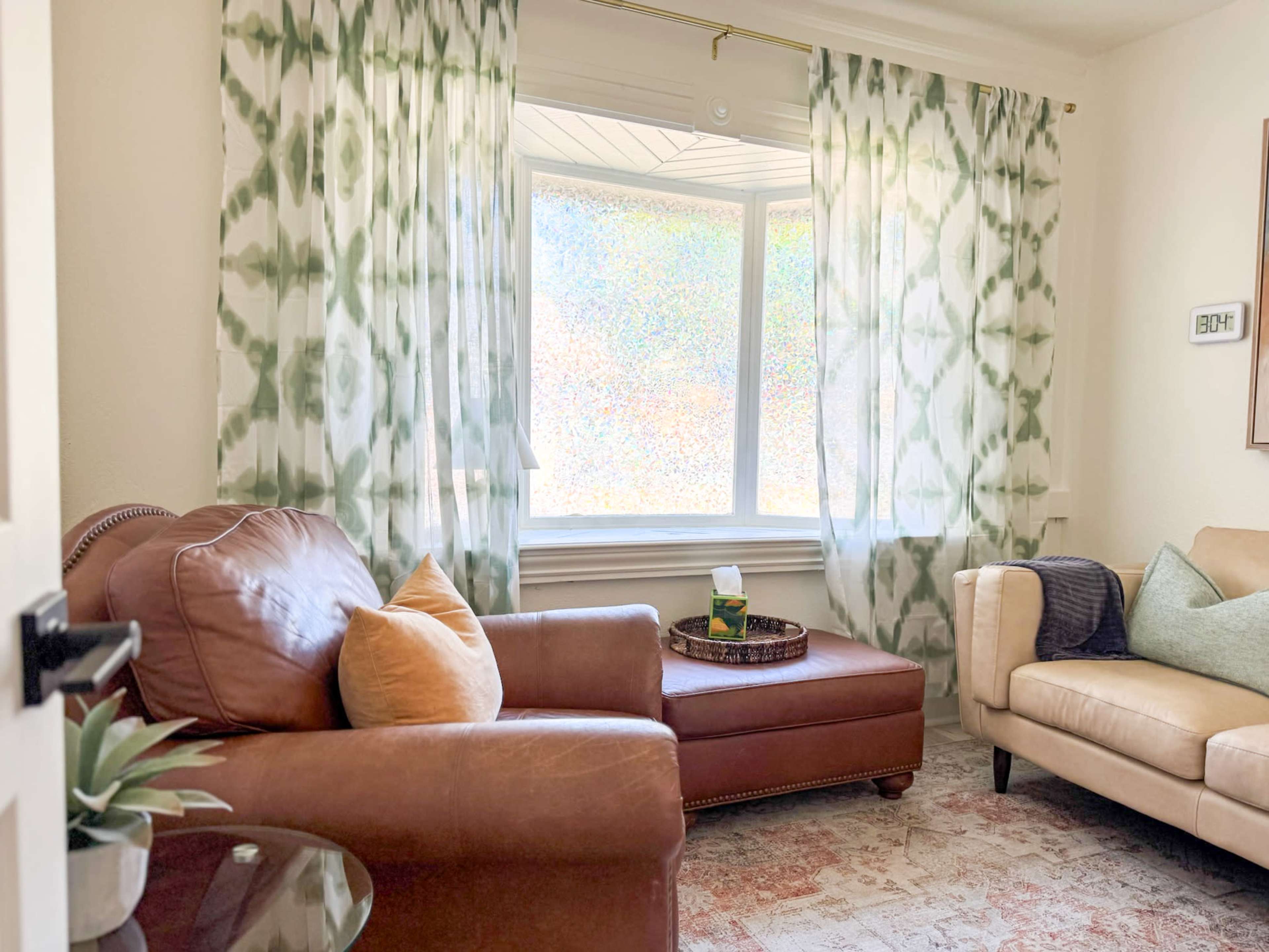 The image shows a cozy living room with a brown leather armchair, a cream-colored sofa, and patterned curtains around a large window.