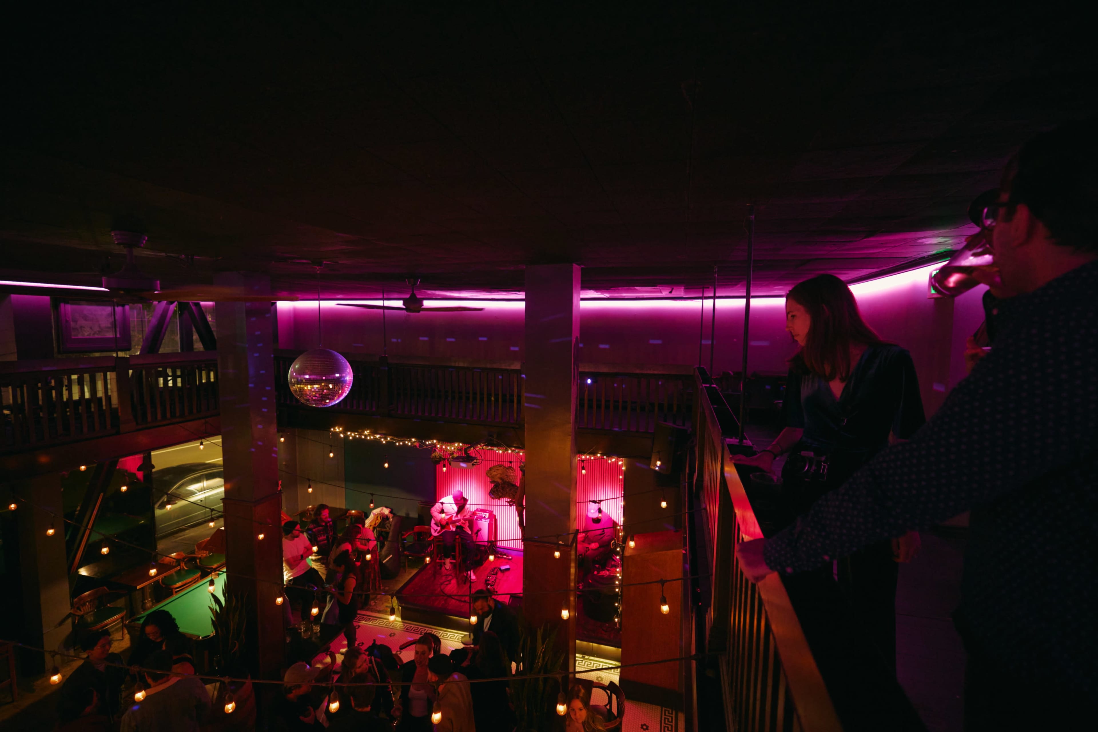 The image shows a lively indoor venue with a band performing on stage, surrounded by an audience enjoying the atmosphere, and colorful lighting illuminating the space.