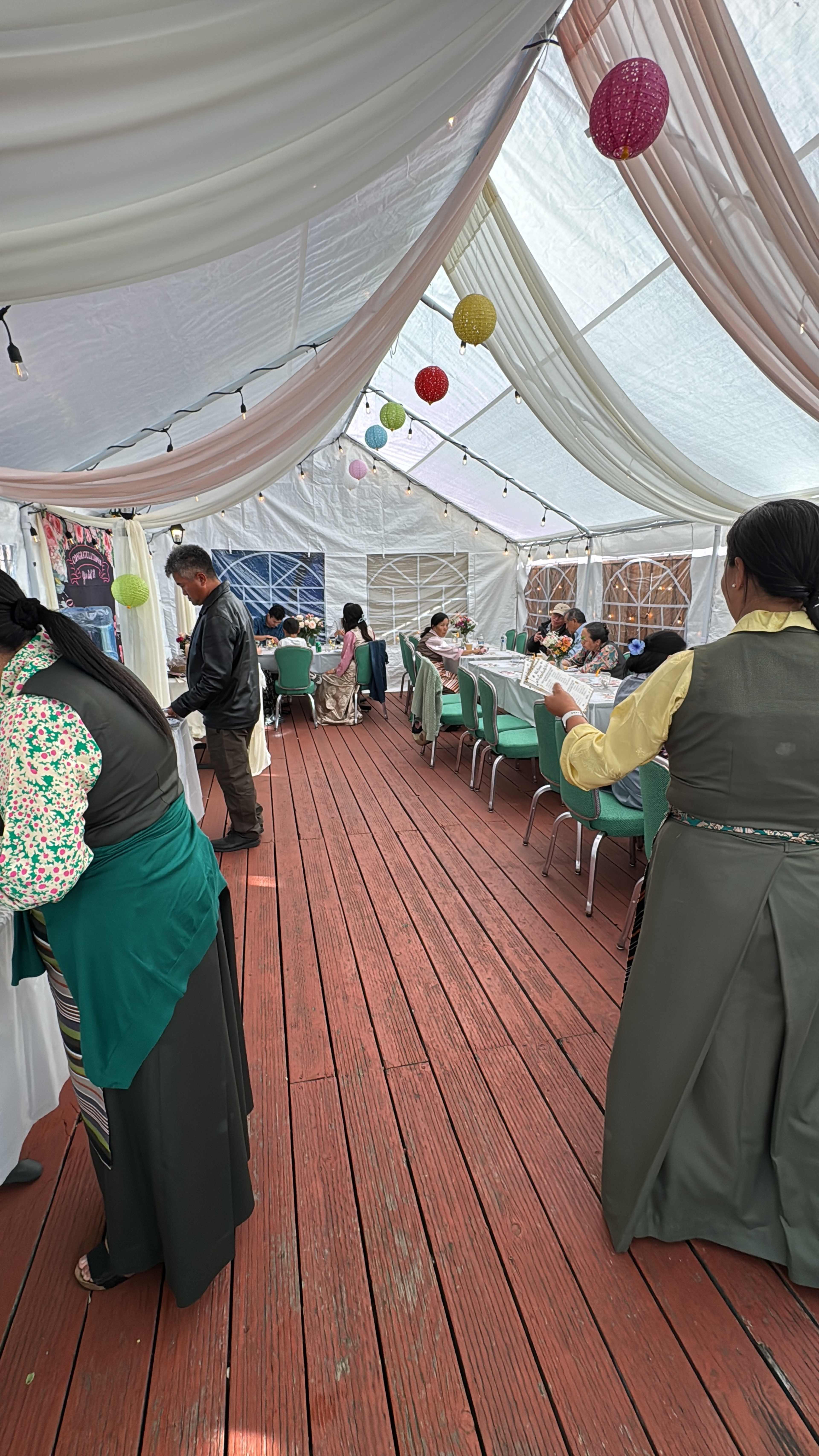 A large, decorated tent features tables set for a gathering, with people interacting and colorful lanterns hanging from the ceiling.