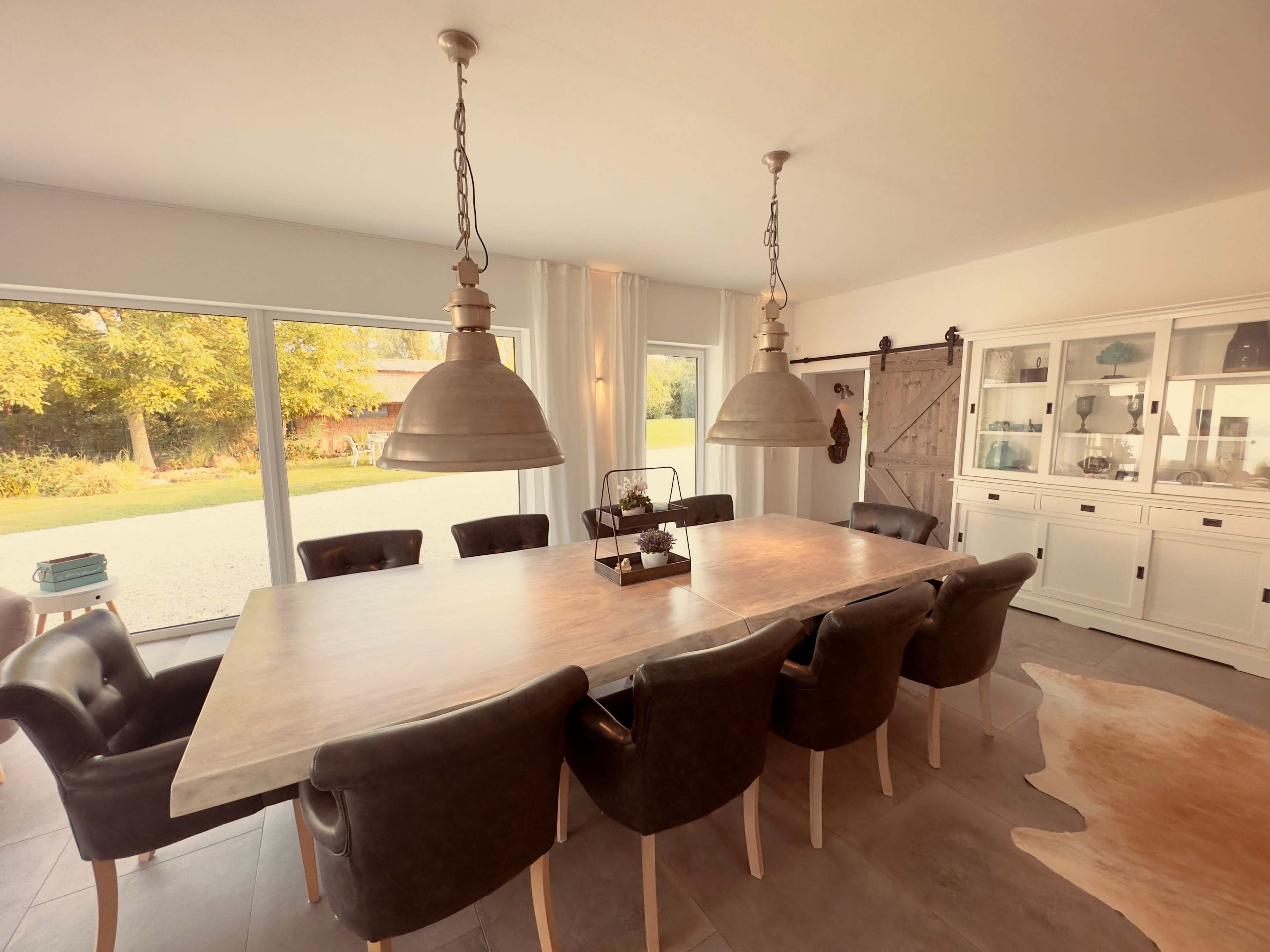 A spacious dining room features a large table surrounded by leather chairs, with large windows providing natural light and a view of the outdoors.