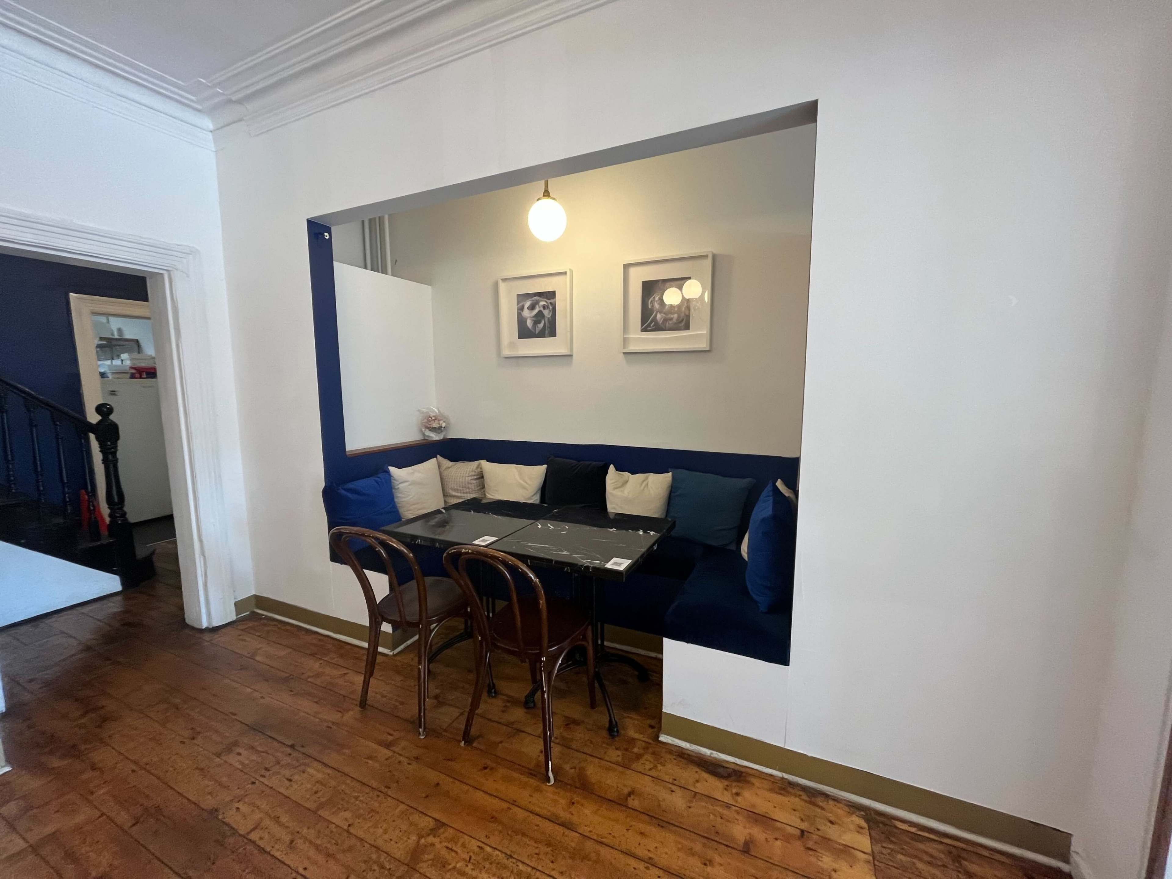 The image shows a small dining nook with a dark table surrounded by blue cushioned seating and three framed pictures on the wall, all set against a white backdrop.