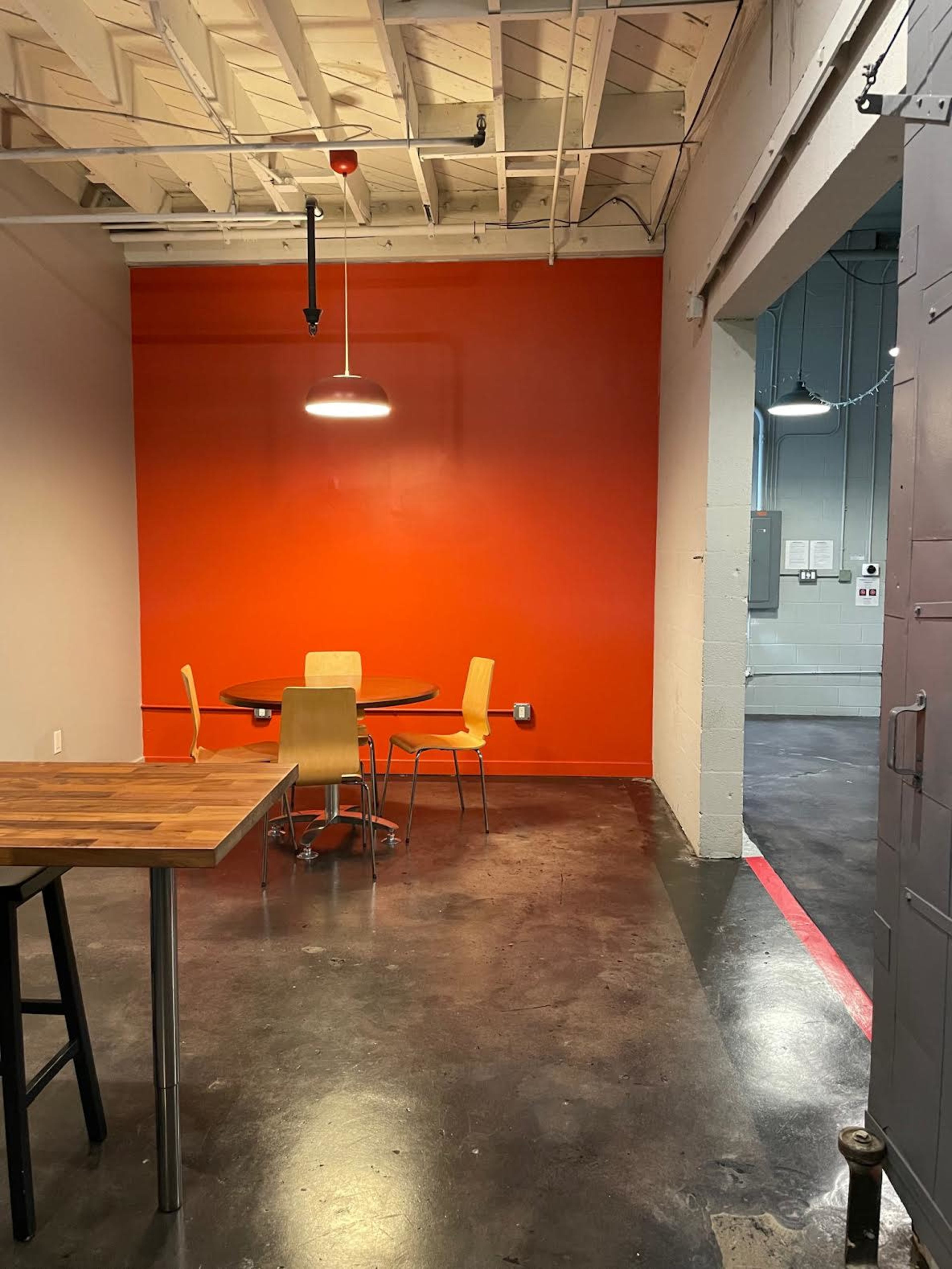 A small dining area with a round table and four chairs set against a bright orange wall, with a ceiling light hanging above.