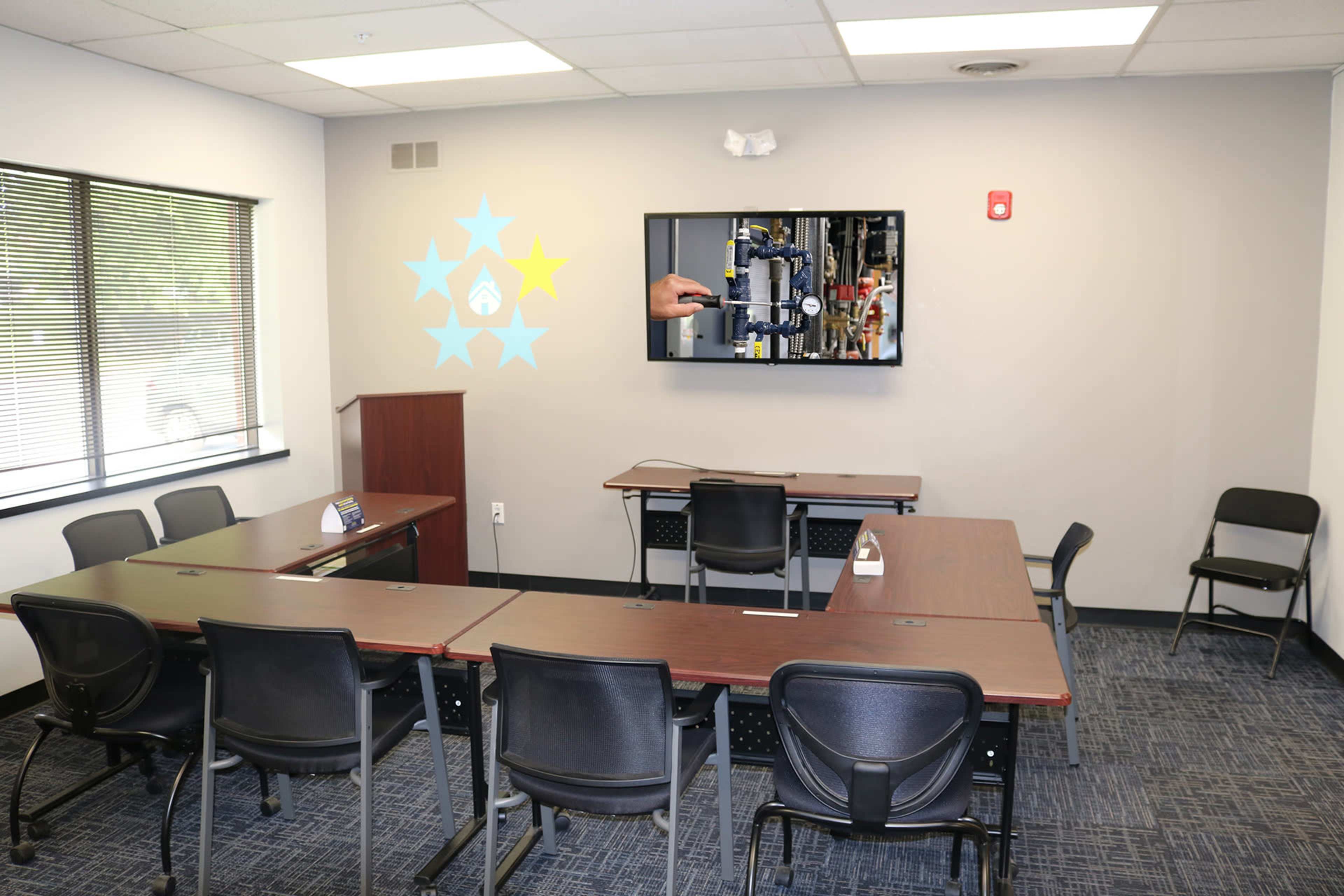 A conference room equipped with rectangular tables, chairs, and a wall-mounted screen displaying a hand using equipment.